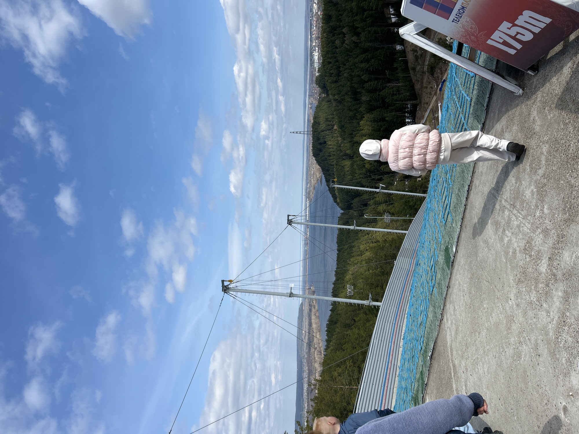 The image captures a panoramic view from the top of a ski jump structure, likely in the Прикамский (Prikamsky) region of Russia, on a bright, clear day with scattered white clouds. The perspective looks out over a vast landscape.
In the foreground, on a textured concrete platform, stands a person dressed in a light pink puffer vest, white hat, and light-colored pants, facing away from the camera and gazing out at the distant scenery. To their right, a large sign is partially visible, displaying "ТЕЛЕКОМ ПАР" and prominently featuring "175m" in white text against a red background. Part of another person, clad in a grey long-sleeved top, is visible in the bottom left corner, seemingly sitting or leaning.
The ski jump's descent is visible to the left of the platform, covered with a blue mesh or matting typical for summer use. The structure includes prominent metal support towers and cables extending across the view. Below the jump, a dense coniferous forest covers the rolling hills. In the mid-distance, a wide body of water, possibly a river or lake, stretches horizontally, reflecting the sky and showing signs of partial ice cover or distant shores. Across the water, a distant city or townscape is discernible on the horizon, blending with the landscape.
The scene depicts an observation or sightseeing moment from a high vantage point, emphasizing the scale of the ski jump and the expansive natural and urban environment. The sunny weather and bright blue sky suggest daytime.