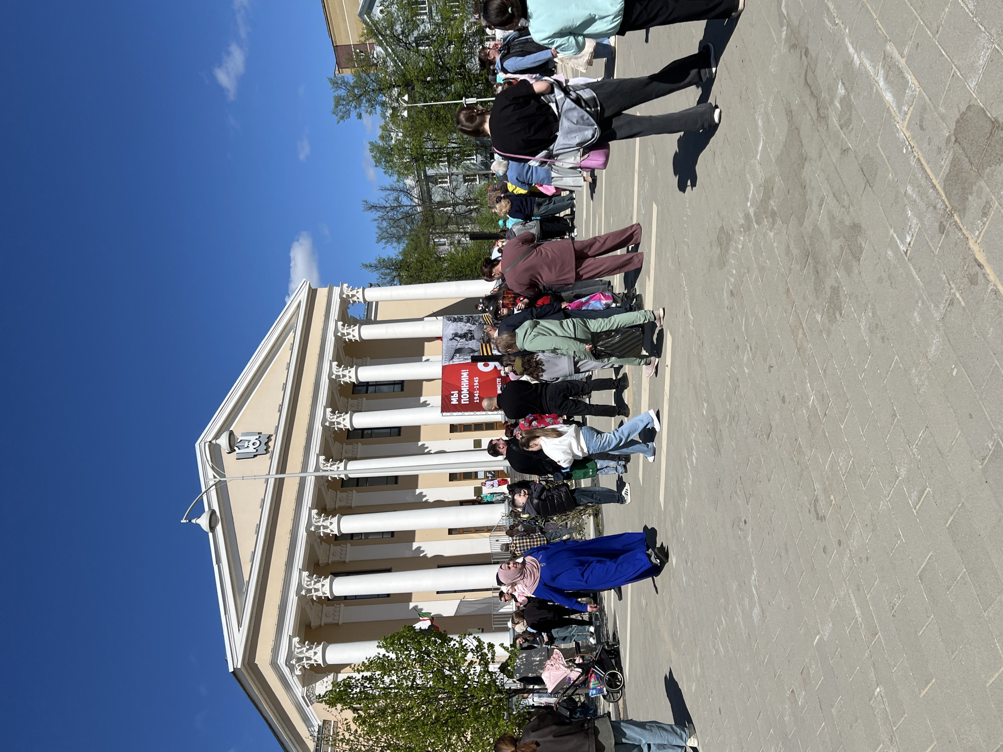 The image depicts a scene in Kazan, Russia, likely near a building with prominent neoclassical architecture, evidenced by its white columns and ornate facade. A group of people are gathered in the paved area in front of the building. A red banner with white text and a black and white image, potentially commemorating an event related to the years 1941-1945, is prominently displayed on the building. The sky is clear and blue, indicating a sunny day. The people vary in age and clothing styles, some carrying bags and wearing casual attire. A few trees provide some greenery around the scene.