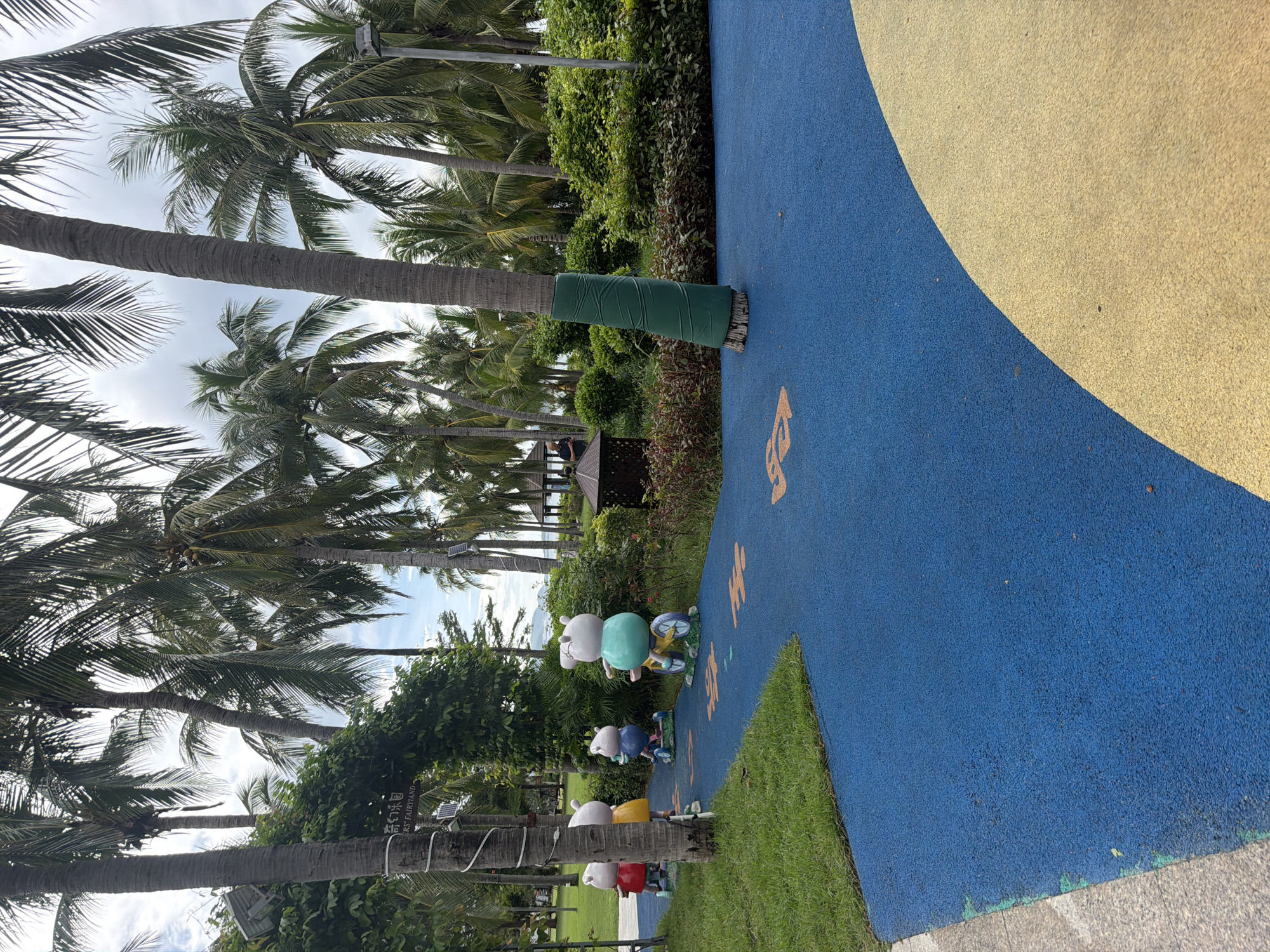 The image depicts a park scene in the Jiyang District of China, with lush palm trees dominating the background under a cloudy sky. In the foreground, a vibrant blue rubberized path winds through the park, bordered by green grass on the left and a section of yellow paving on the right. The path is decorated with orange paw prints. Along the path, several cartoonish animal statues are visible. The first statue, on the far left, is a piglet wearing a red dress. To its right is another piglet in a yellow dress, followed by a piglet in a blue outfit on a scooter. The furthest statue on the right is a piglet in a green top riding a yellow bicycle. In the midground, a small, dark brown wooden gazebo with a pointed roof is situated amidst dense green foliage. The base of a prominent palm tree in the mid-right of the image is wrapped in green material. Further back, more palm trees and greenery suggest a tropical or subtropical environment. The scene appears to be a public recreational area, possibly a playground or a themed park for children.