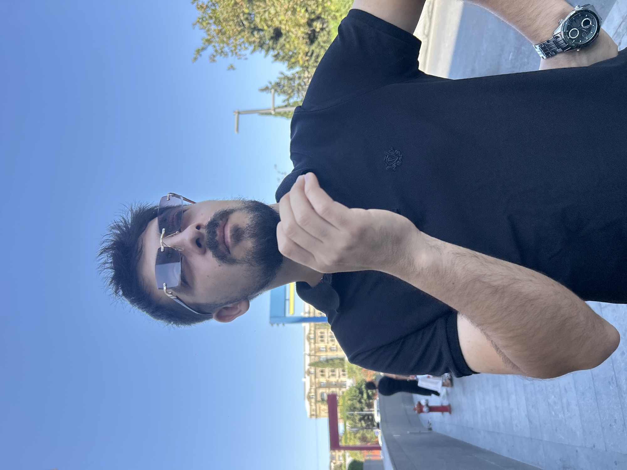 A man with a beard and sunglasses is looking up and to the right, with his hand near his chin. He is wearing a black polo shirt and a watch on his left wrist. In the background, there are buildings, trees, and a blue sky. The man appears to be outdoors in a city. The buildings in the background suggest the location is in Baku, Azerbaijan, possibly near the Fountain Square or Nizami Street area, given the architectural style and urban setting. There are also a few other people visible in the distance, walking along a sidewalk. The lighting suggests it is daytime.