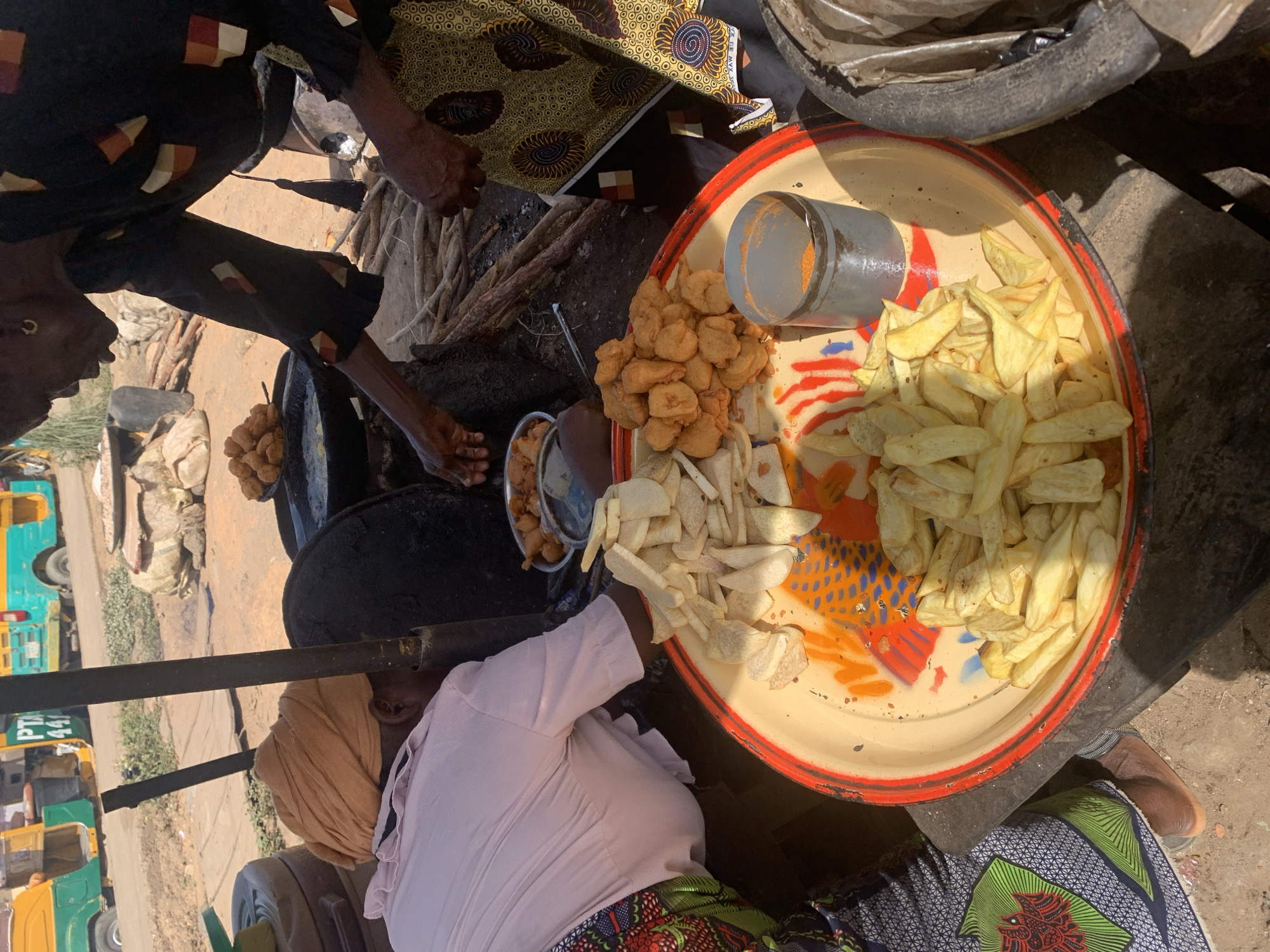 An outdoor scene in Rafin Par, Nigeria, depicts a food vendor preparing street food. The vendor, wearing a light pink shirt and a patterned skirt, is seated next to a large tray containing fried yams, akara (bean cakes), and sliced yams. A grey container likely holding spices sits on the tray. An individual in a patterned top reaches towards a pot, possibly to retrieve more food. A frying setup sits to the left. Auto rickshaws are visible in the background. The setting appears to be a dusty roadside.