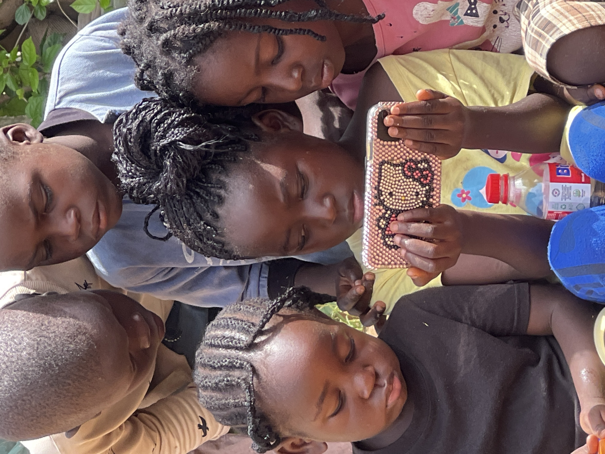 In Jos, Nigeria, a group of African children are gathered around a smartphone. The children, ranging in age, are focused on the device, which is held by two girls in the foreground. The phone has a pink, sparkly case with a cartoon character design. The children are dressed in casual clothing. The environment appears to be outdoors, with hints of greenery in the background, suggesting a possibly rural or less urban setting. There is a bottle of water with a red cap on the ground next to the children.