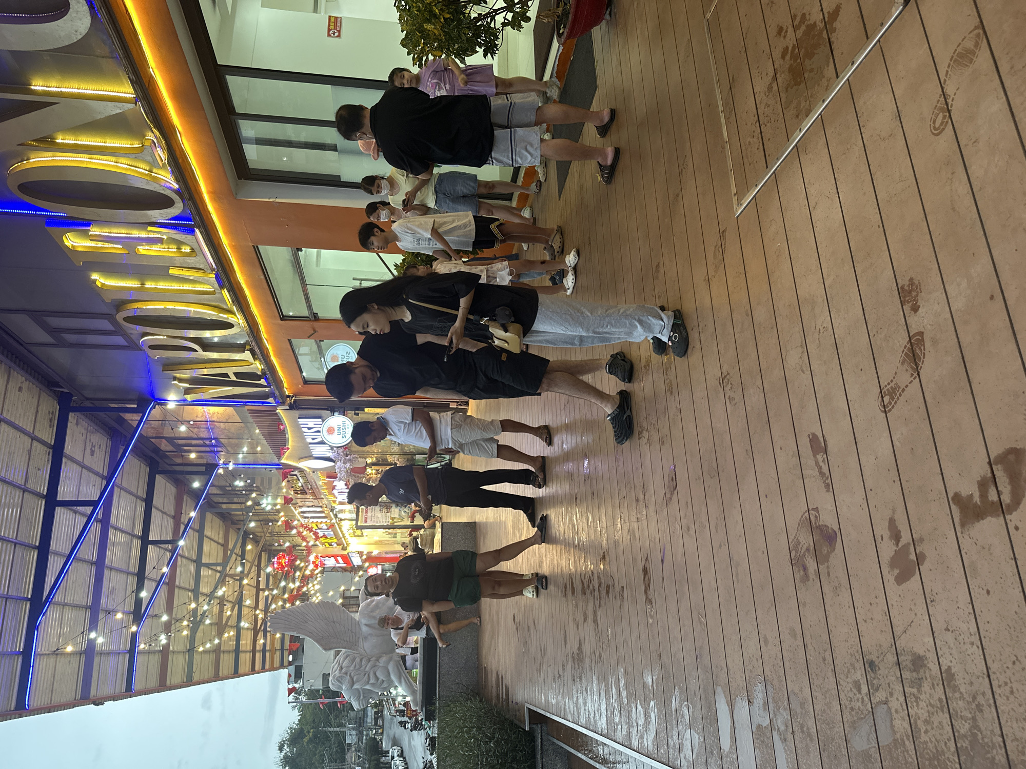 A long line of people stand on a wooden boardwalk outside a brightly lit establishment, the boardwalk marked with water stains and footprints, presumably from rain. They appear to be waiting to enter a restaurant or shop that has signage in a non-English language. In the background, lights and an angel statue mark what appears to be a pedestrian thoroughfare, consistent with the urban location of Bắc Nha Trang Ward, Vietnam.