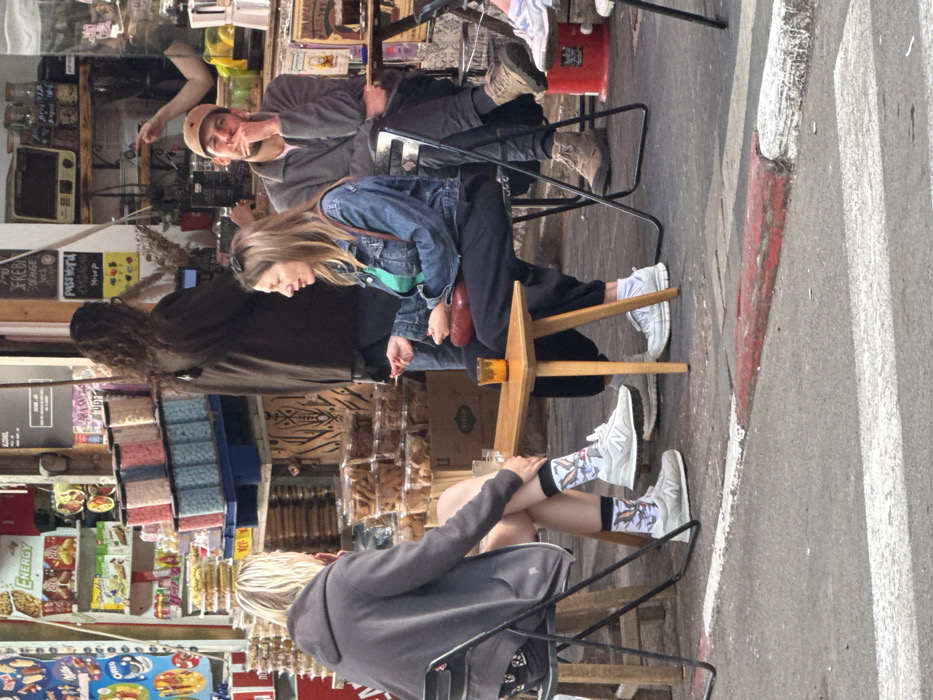 The image depicts an outdoor cafe scene on a city street, characteristic of Tel-Aviv, Israel, given the visible Hebrew text and urban environment. In the foreground, an asphalt street features a white crosswalk line and a red-and-white painted curb.
Three individuals are seated at small, light wooden tables on the sidewalk. On the left, a person with short, light-colored hair, wearing a grey hooded sweatshirt, dark shorts, patterned socks, and light grey New Balance sneakers, sits with their back mostly to the viewer, facing right. Their left leg is crossed over their right knee, and a clear glass containing an amber liquid rests on the table in front of them.
Opposite this person, a woman with long, light brown hair, dressed in a denim jacket over a green top and dark track pants, is seated. She holds a lit cigarette in her right hand and is looking towards the left, engaged in conversation. A red shoulder bag is visible on her lap, and she wears white socks and light grey sneakers.
To the right of the woman, a man with a light brown baseball cap, a grey collared shirt or jacket, and dark pants is seated, partially obscured. He has a slight beard and is resting his chin on his left hand, looking towards the left. He wears light brown hiking-style boots.
Behind these individuals is a shop or cafe storefront. Its left side displays shelves stocked with packaged snacks, including "ENERGY" bars and "Oreo" cookies. Below these, numerous clear plastic containers filled with different colored candies or nuts are stacked on a blue pallet. Further right, more plastic containers of baked goods or cookies are stacked. The interior of the shop features a counter with a coffee machine, various jars, and a handwritten "PASSWORD" sign. In the background, a person with long, dark, curly hair in a dark jacket stands inside the shop, facing away from the viewer. Another arm and hand are visible reaching over a counter on the far right. The scene is illuminated by natural daylight.