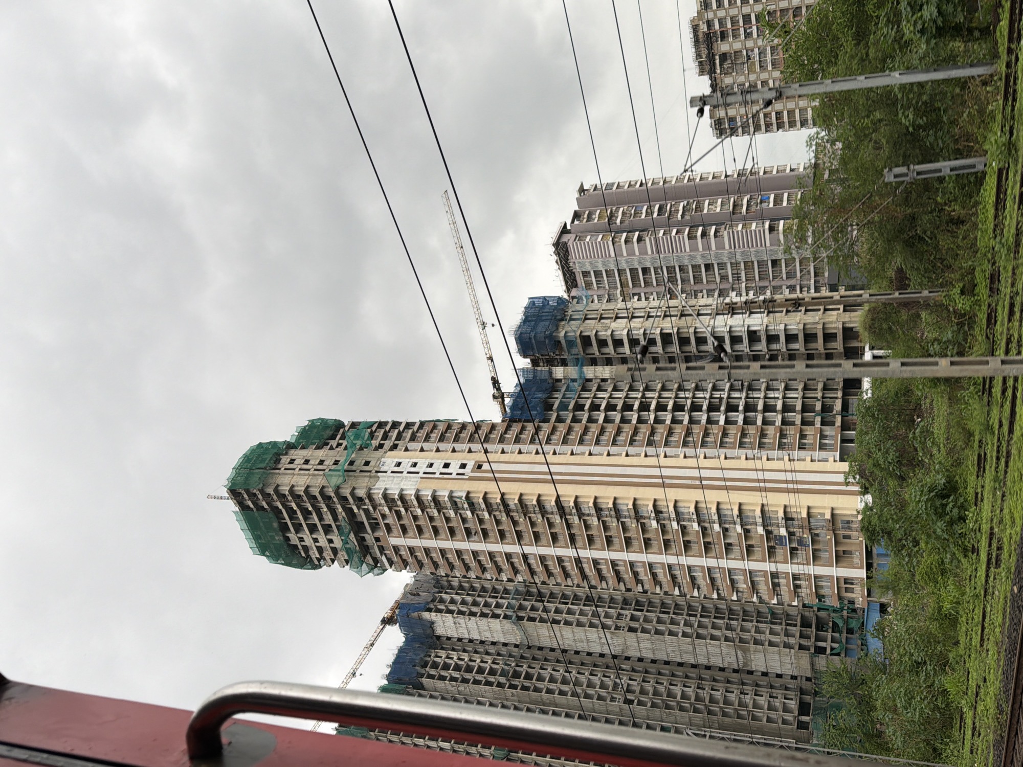 The image depicts a view of a partially constructed high-rise building in Mira-Bhayander, India, against a cloudy sky. The building, covered with green netting, appears to be in the midst of construction, with cranes visible in the background. Several other tall buildings, some also under construction, are present in the vicinity. The foreground shows railway tracks and lush green vegetation, suggesting the photo was taken from a moving train. The overall scene conveys urban development in progress.