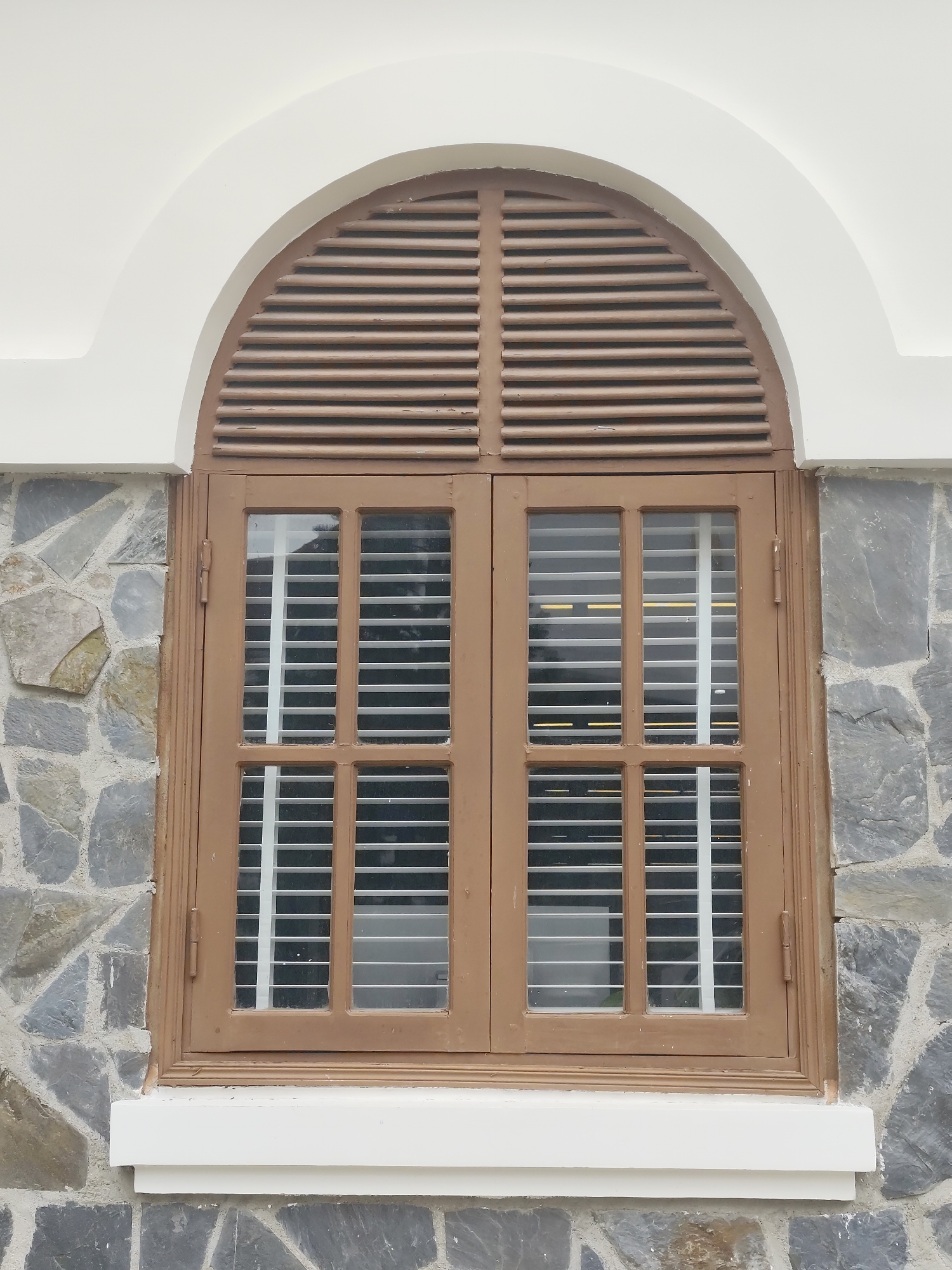 The image shows a close-up of a vintage arched window with wooden shutters and a stone wall in Lâm Viên - Đà Lạt, Vietnam. The window is framed by a white arched structure above and a white sill below. The upper part of the window features horizontal wooden louvers, while the lower part consists of two double-paned windows with white blinds. The wall is constructed from irregular gray and tan stones. The lighting suggests it is daytime. There are no people or animals visible in the frame. The scene is architectural and appears to be an exterior view of a building.