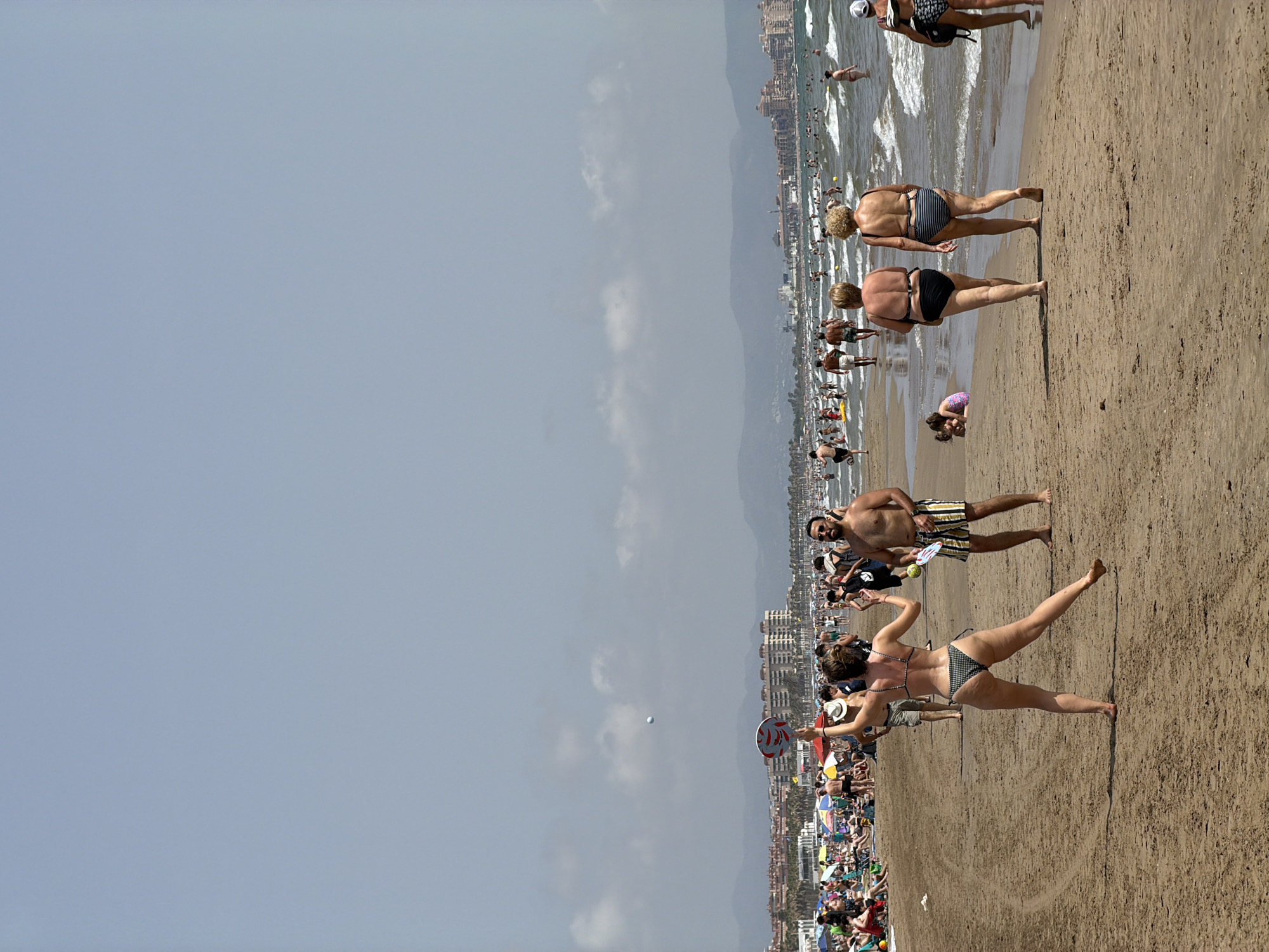 A wide shot captures a crowded beach on a sunny day. In the foreground, a woman in a black and white checkered bikini is playing beach paddleball, with a red and white paddle raised. Behind her, a man in striped shorts holds a yellow and white paddle. Further back, two women in bikinis walk away from the camera towards the water. The ocean is filled with swimmers, and beyond the beach, a city skyline with buildings and palm trees stretches along the coast. The sky is pale blue with scattered clouds.