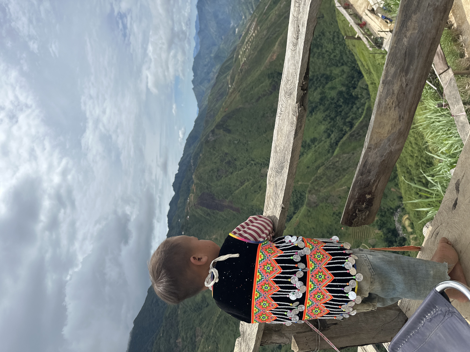 A young child, viewed from behind, stands on a wooden platform, looking out at a mountainous landscape. The child wears a traditional, intricately decorated vest with colorful geometric patterns and hanging silver coins, and simple denim pants. The vest is black with a band of vibrant embroidery in pink, yellow, and green, accented with small white beads. The child's hair is short and light brown.
The background reveals a vast expanse of rolling green mountains, terraced fields, and a cloudy sky. The terrain suggests a rural, mountainous region, consistent with the description of Ta Xua, Vietnam, known for its stunning natural beauty and tea plantations. The wooden platform appears rustic, with rough-hewn planks and a simple wooden railing. A dark chair is partially visible in the foreground to the left. The overall scene captures a moment of quiet contemplation against a breathtaking natural backdrop.