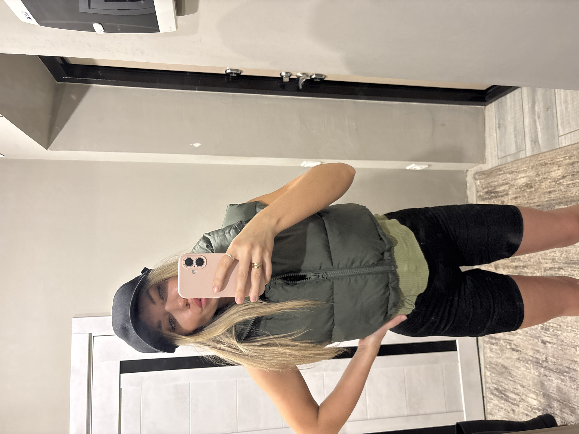 An individual is depicted taking a mirror selfie in what appears to be an indoor hallway or entrance area in Votkinsk, Russia. The female subject, with long blonde hair and wearing a black baseball cap, is seen from the waist up. She is dressed in an olive green cropped puffer vest over a light green top, paired with black cycling shorts. Her left hand holds a light pink smartphone, which partially obscures her face, with her lips slightly pursed. Two gold-colored rings are visible on her left ring finger. Her right hand rests on her hip.
The background features a light gray wall with minor scuffs and a white door with dark vertical accents on the left. On the right, a dark-framed door with visible silver hardware (handle and locks) is present. Above and to the right of the door, a white and black wall-mounted device, possibly an intercom or security panel with the text "ZAI" on its upper left, is affixed to the wall. The floor consists of light gray tiles, with a portion of a textured light-colored rug visible on the left foreground. A black boot or shoe is partially visible on the far left.
