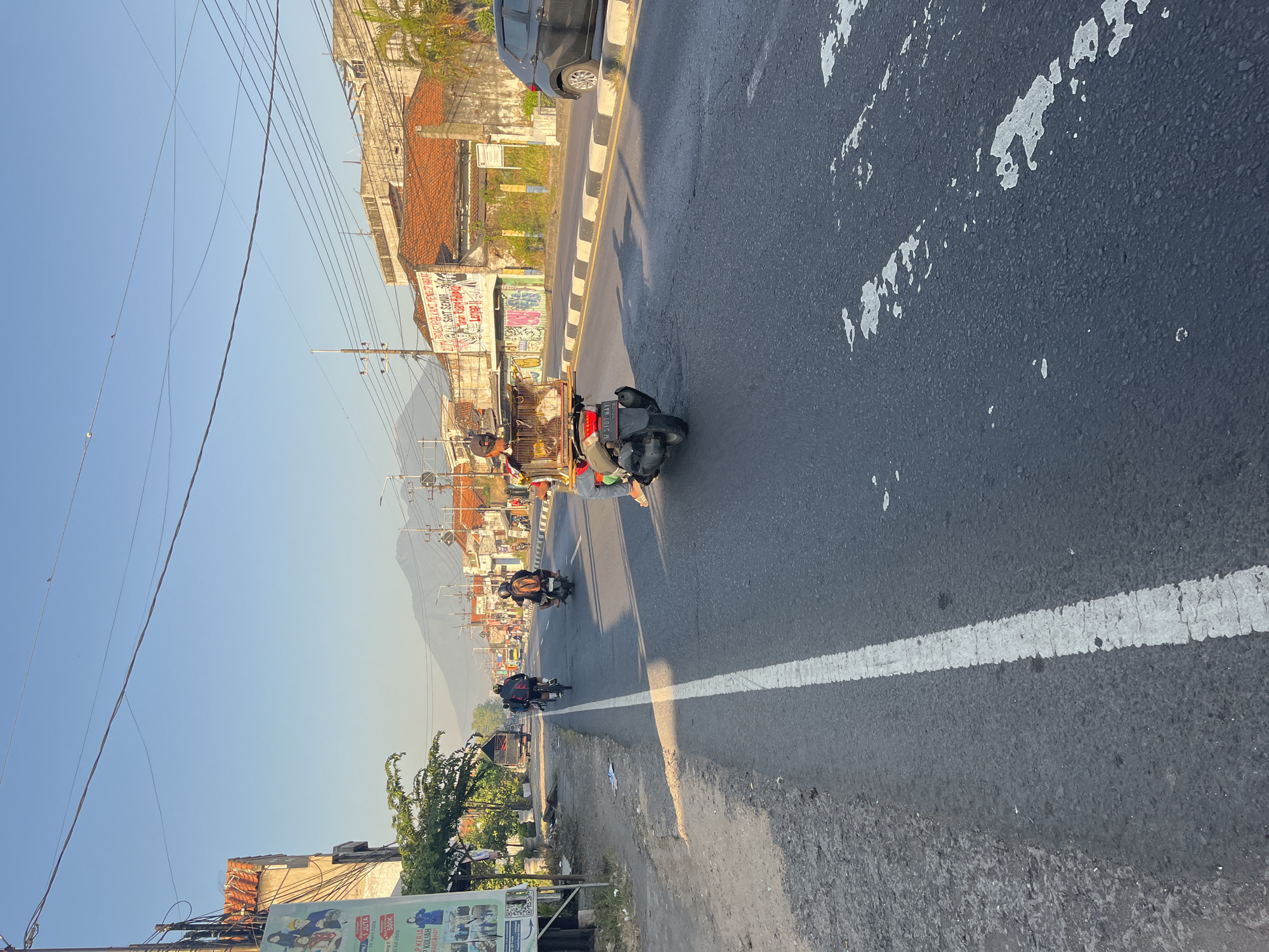 In the city of Gedang, Indonesia, a sunny day illuminates a bustling road. A person on a scooter transports a birdcage, its feathered occupants visible within. Several other vehicles, including motorbikes and a truck, travel on the asphalt road, lined with buildings and trees. In the distance, a majestic mountain looms under a clear blue sky. A billboard stands on the left side of the road, its message obscured by the angle. This scene captures a slice of daily life on a busy Indonesian street.