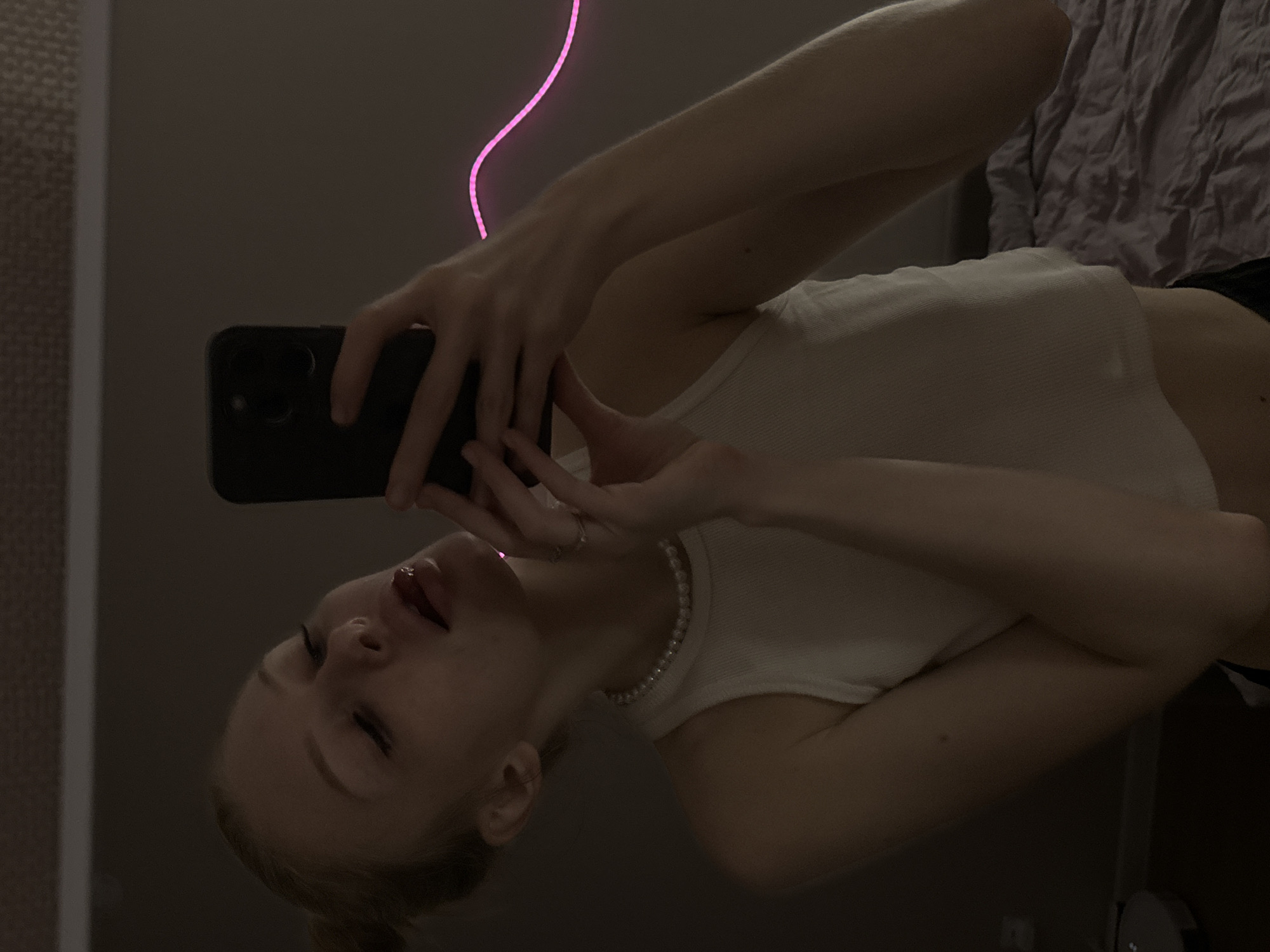 An indoor scene, presumably a bedroom, is depicted in a mirror selfie taken in the city of Perm, Russia. A fair-skinned female with blonde hair styled in a bun is the central subject, captured from the chest up. She wears a white ribbed tank top and a pearl necklace. Her head is tilted slightly back and to the left, with her eyes mostly closed and lips parted, displaying a glossy, light-colored product. She holds a black smartphone with her right hand, which has rings on the fingers, aimed towards the mirror to take the photo. The room is dimly lit, with a distinctive wavy pink neon light strip affixed to the wall in the upper right background. A purple, unmade duvet on a bed is partially visible on the right, and a light-colored circular robot vacuum cleaner rests on the dark floor in the lower left corner.