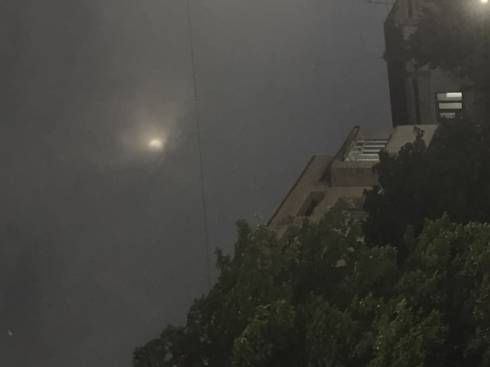 This is a nighttime or dusk photograph taken in Tashkent, Uzbekistan, capturing a building partially obscured by trees against a dark, cloudy sky. A bright, hazy light source, likely the moon or a streetlamp, is visible in the upper left quadrant, diffused by the clouds. The building on the right appears to be multi-story with windows that are lit from within, suggesting occupancy. The foreground is dominated by the dark foliage of trees, adding a sense of depth and natural elements to the urban setting. The overall mood is atmospheric and somewhat mysterious due to the low light conditions and the obscured nature of the scene. No people are clearly visible, and no text is discernible within the image.