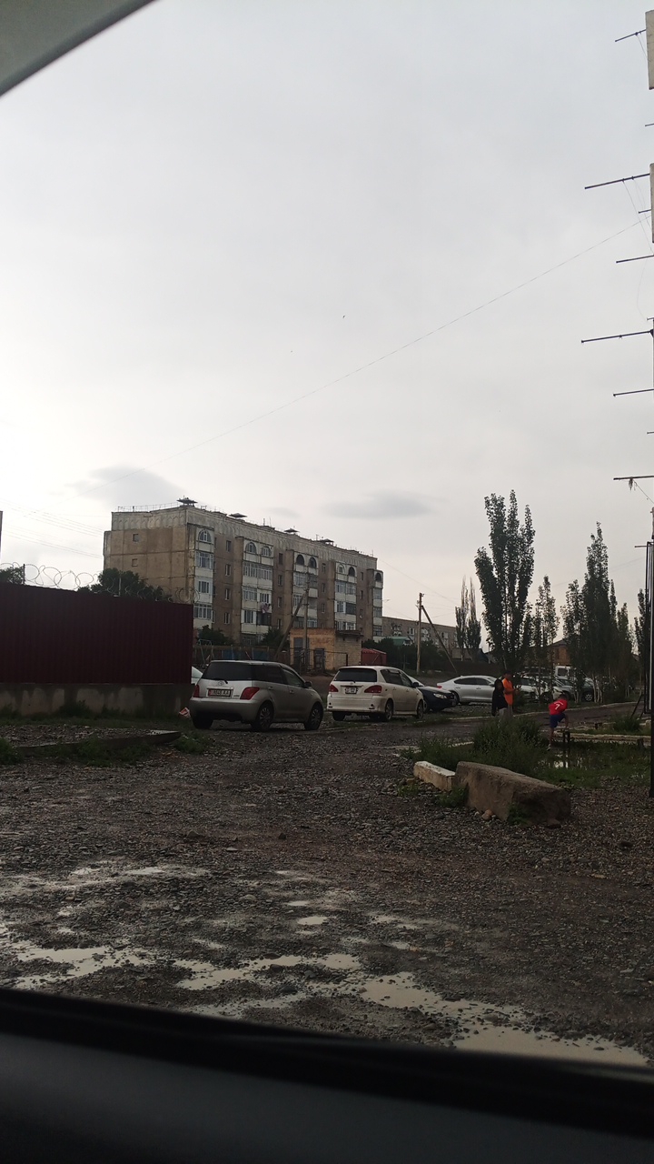 A view of a street in Balykchy, Kyrgyzstan, shows a multi-story apartment building in the background with a light gray, overcast sky. Several cars are parked on the gravelly, wet street in front of the building. A dark red fence runs along the left side of the image. Tall, slender trees line the right side of the street. The ground is covered in a mix of gravel and puddles of water, suggesting recent rain. In the distance, people can be seen near the trees.
