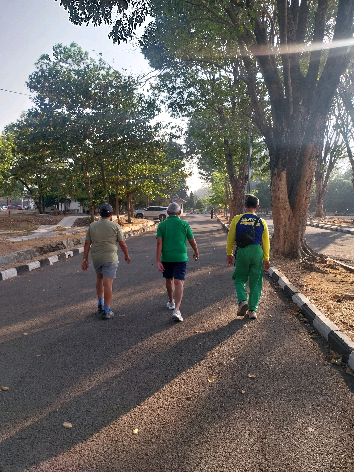Three men are walking on an asphalt path lined with trees and a curb on the right side. The path appears to be in a park or recreational area. The man on the left wears a light green t-shirt, grey shorts, blue socks, and dark shoes. The man in the middle wears a green t-shirt, blue shorts, and white athletic shoes. The man on the right wears a yellow long-sleeved shirt with blue accents and "DIKLAT FIT" written on the back, green pants, and beige athletic shoes. He also has a black fanny pack around his waist. The sun is shining, casting long shadows across the path. In the background, there are more trees, some buildings, and parked cars. The overall setting suggests a pleasant outdoor environment, possibly within a city park in Cirebon, Indonesia, given the context of the request.