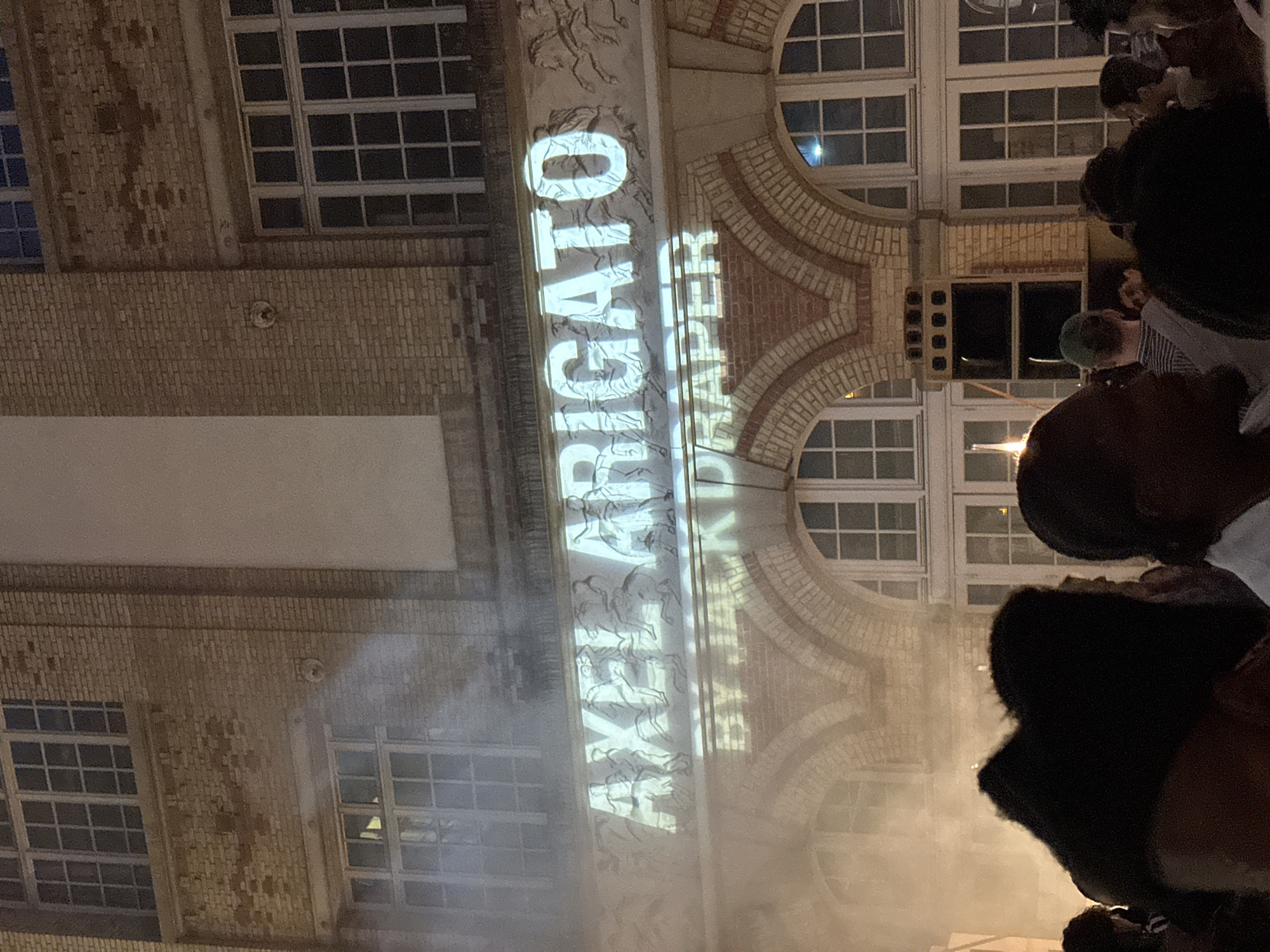 The image captures a nighttime scene in Paris, France, focusing on a building facade with an architectural style featuring brickwork and multiple windows. Projections illuminate the building, displaying text that reads "ARELYKIPAPER" and "BRIGATO." Several individuals are present, appearing to look up at the illuminated building. The overall environment has a slightly smoky or hazy quality, possibly from the projection equipment or atmospheric conditions.