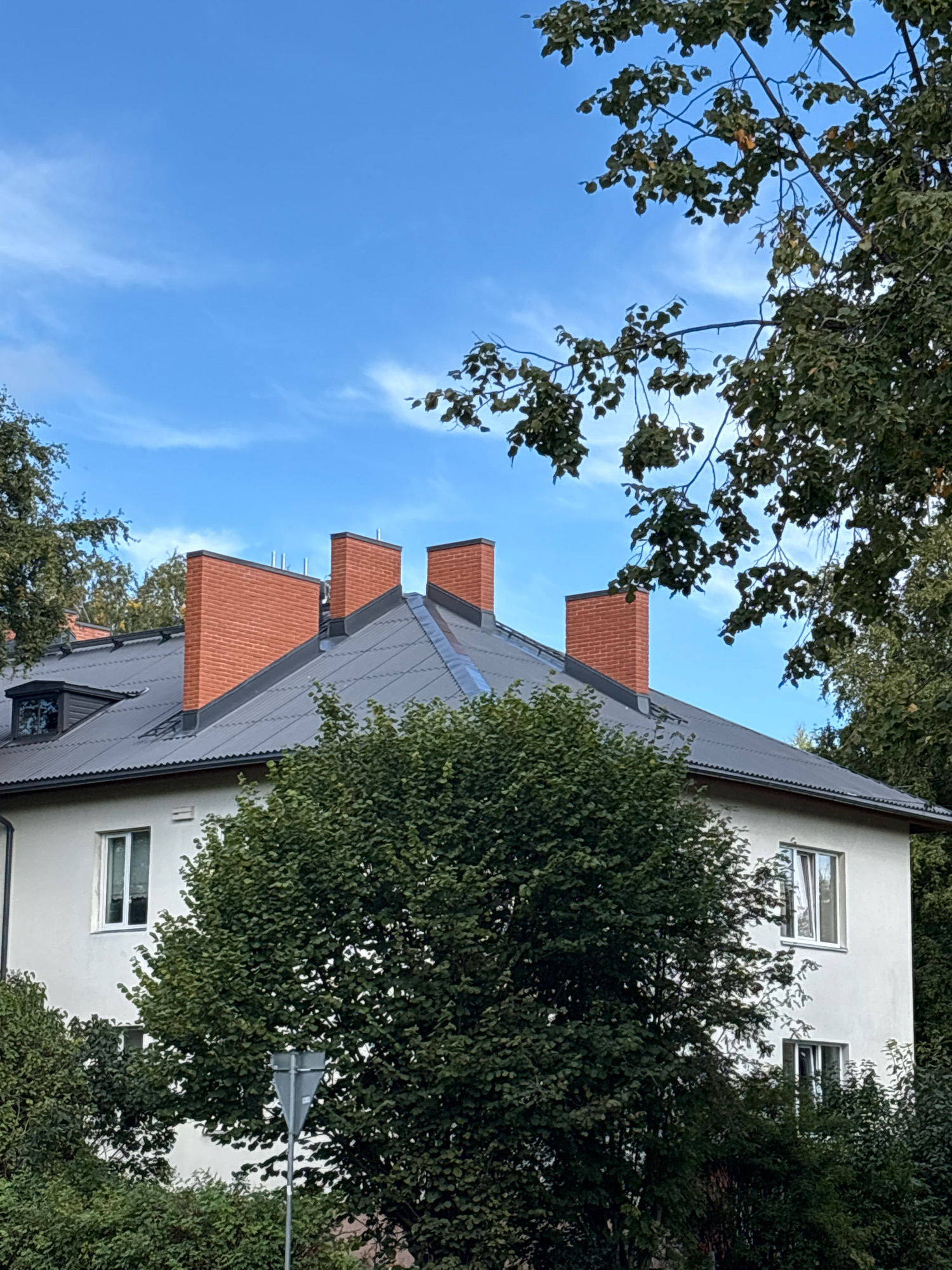 The image shows the roofline of a light-colored building with a dark metal roof. Multiple brick chimneys are visible on the roof. A dormer window is present on the left side of the roof. Trees with green foliage partially obscure the building. The sky is blue with some wispy white clouds. The scene is outdoors, and no people or animals are visible. The image is a photograph taken from a medium distance, focusing on the upper portion of the building and the sky. There is no indication of the specific location in Tallinn, Estonia.