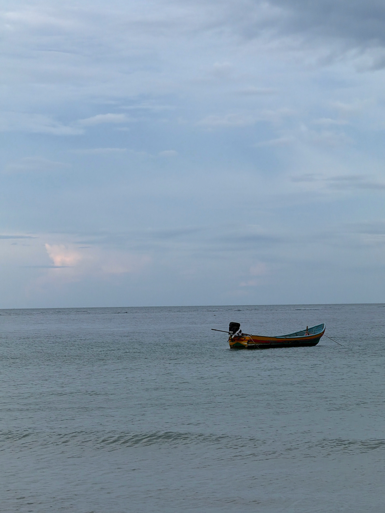 A small, colorful fishing boat with a dark outboard motor is moored on calm, gray water under an overcast sky. The boat, which appears to be made of wood, has orange, yellow, and blue accents. It is anchored by a rope that disappears into the water. The scene is likely a coastal area in the Ko Samui District of Thailand, given the style of the boat and the tropical clouds.