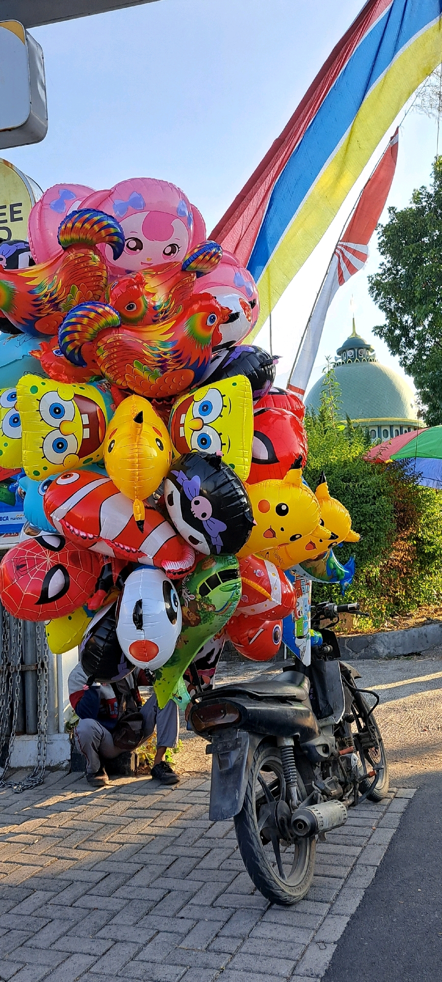 In the city of Prigen, Indonesia, a vendor sits on a brick sidewalk next to a parked motorcycle laden with a colorful assortment of inflatable balloons. The balloons depict various cartoon characters such as SpongeBob SquarePants, Pikachu, Spider-Man, and a clownfish, alongside animal shapes like roosters and a pink character with blue bows. Behind the balloons and motorcycle, a lush green hedge is visible, with a domed building featuring a green roof and ornate top rising in the distance. A series of vertical, multi-colored banners, red, yellow, and blue, are strung diagonally across the sky. The vendor, wearing a jacket and grey pants, is crouched down, partially obscured by the balloons. The scene is illuminated by natural sunlight, casting shadows on the brick pavement.