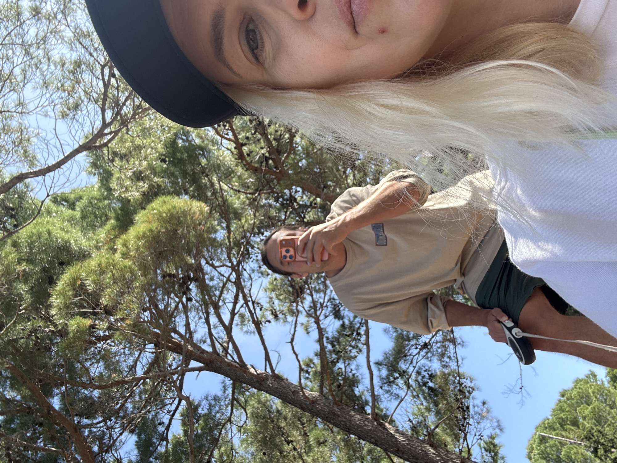 A photograph taken in Petrovac centar, Montenegro, shows a person holding a retractable dog leash and taking a selfie with their smartphone, positioned below another individual who is partially visible at the top of the frame. Both are outdoors under the shade of trees. The person holding the leash is wearing a light brown t-shirt and green shorts. The upper person has blonde hair and is wearing a black hat and white shirt. The background consists of trees and a clear sky.