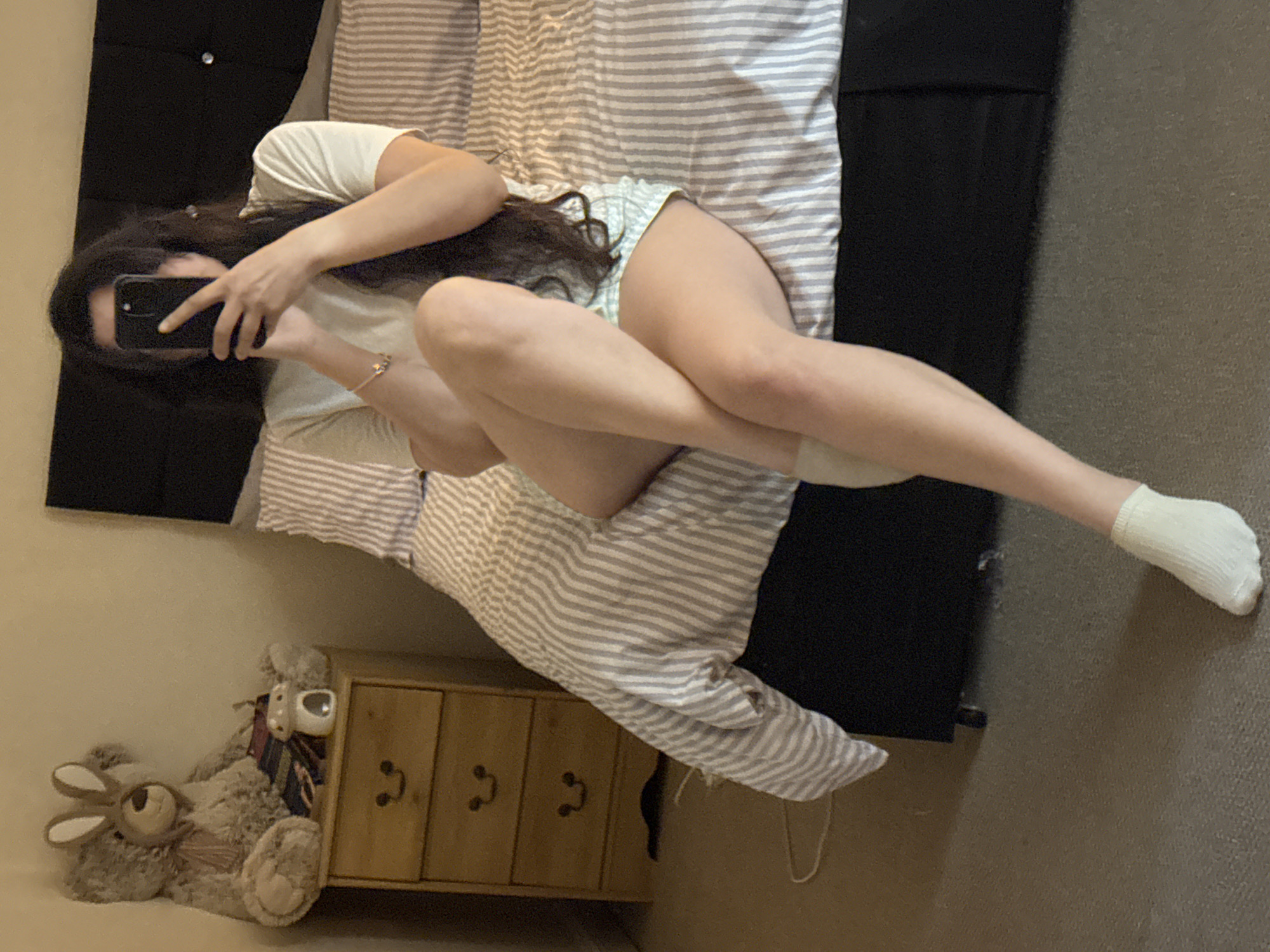 A woman is lying on a bed with a gray and white striped duvet. She is holding a black smartphone, presumably taking a picture of herself. She is wearing a white t-shirt and patterned shorts. Her legs are crossed. One leg is wearing a white sock. Next to the bed is a small wooden chest of drawers with three drawers and metal handles. A stuffed rabbit and dog are lying next to the chest of drawers. The headboard of the bed is black. The background includes a beige wall and carpet. The location is not apparent from the image and it is impossible to confirm it is Great Yarmouth.