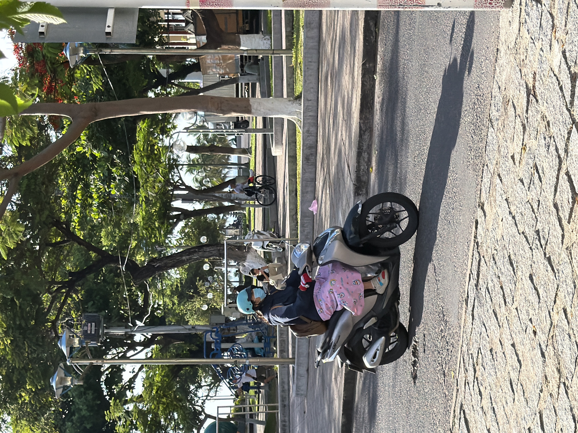 A woman wearing a light blue helmet, a face mask, a dark jacket, and a pink patterned skirt rides a silver scooter on a paved road. She is wearing white sneakers with red and blue accents. The scooter has a black backpack strapped to its back. The woman is riding on the right side of the road, casting a long shadow to her left. The road surface is asphalt with a cobblestone sidewalk to the left. In the background, there are trees with green foliage and some with red flowers. There are street lamps and some playground equipment. Further in the background, there are people walking and sitting, and other scooters and a bicycle. The scene appears to be in a park or public area within Nha Trang, Vietnam, likely along the coast given the presence of palm trees in the distance.