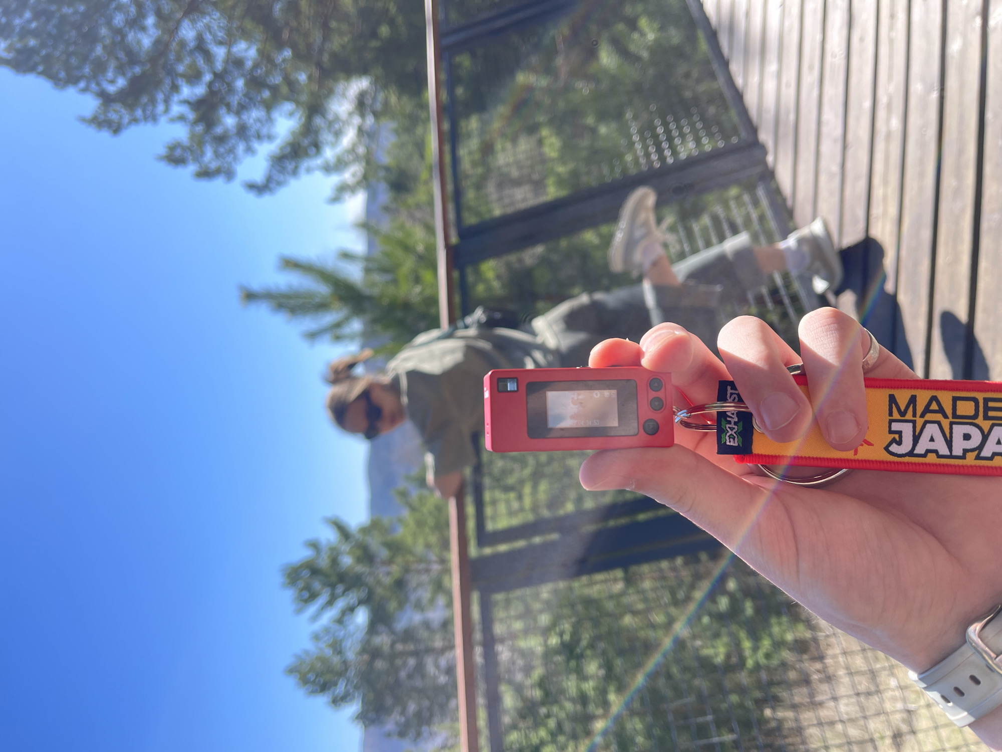 A person stands on a wooden observation deck in Krasnoyarsk, Russia, holding a red digital camera with a "Made in Japan" keychain. The person on the observation deck wears olive-green pants, white shoes and a black backpack. A second person wearing sunglasses and an olive-green top stands behind the deck railing, looking at mountains. The sky is blue, with scattered trees in the background.
