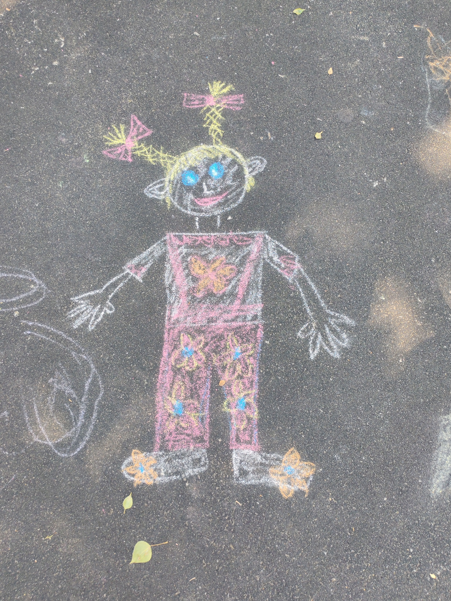 The image shows a chalk drawing of a child-like figure on an asphalt surface, likely a playground or sidewalk. The figure has a round head with large blue eyes, a smiling mouth, and yellow hair styled with pink bows. It is wearing a pink outfit with yellow and blue floral patterns and a butterfly design on the chest. The figure has outstretched arms with detailed hands and is standing with feet adorned with yellow star-like shapes. The drawing is in color, using chalk pastels.
The setting appears to be outdoors, with some fallen leaves scattered on the asphalt, suggesting it might be autumn. The lighting suggests daytime. The overall mood of the drawing is playful and innocent. There is no visible text in the image. The presence of chalk art on the ground indicates a recreational activity, possibly by children.