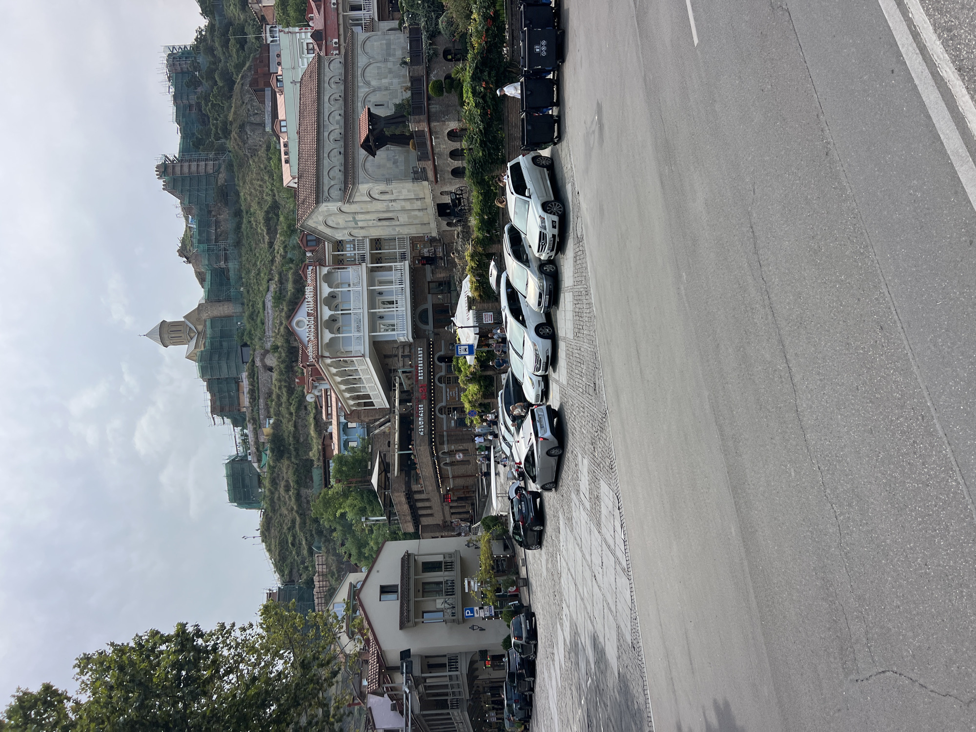 The image shows a street scene in the Old Town of Tbilisi, Georgia. In the foreground, a paved street transitions to a cobblestone area with parked cars and several large black trash bins. Several vehicles, including a white Mercedes SUV, a silver Toyota SUV, and a silver Toyota sedan, are parked along the side. A person in a white hat is walking next to the trash bins. In the mid-ground, there are buildings with traditional balconies, cafes, and restaurants. Signs indicate "Restaurant" and "Cafe." A few pedestrians are visible walking in this area. In the background, a hill is covered with buildings and scaffolding, suggesting ongoing construction or renovation. A prominent church with a dome is situated on the hilltop, and a fortress-like structure is also visible, also covered in scaffolding. The sky is overcast.