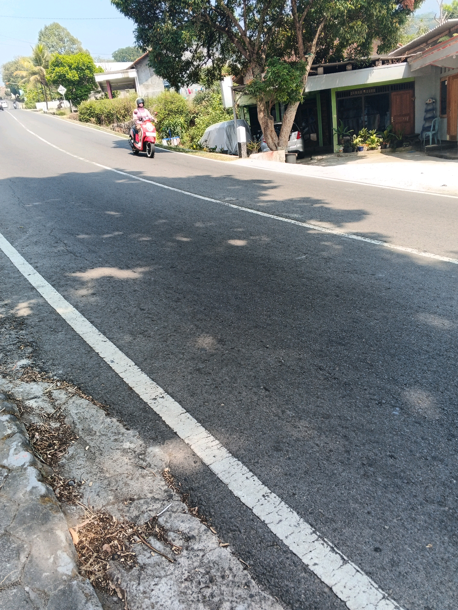 A person wearing a pink shirt and black helmet rides a red scooter on a paved road with a white line marking the lane. The scooter is in motion. The road slopes upwards. To the right of the road, there are buildings with storefronts, trees, and vegetation. A car is parked in front of one of the buildings. The sky is clear and blue. The image appears to be taken during the daytime in a semi-urban or rural area. The location is identified as Pakopen, Indonesia.