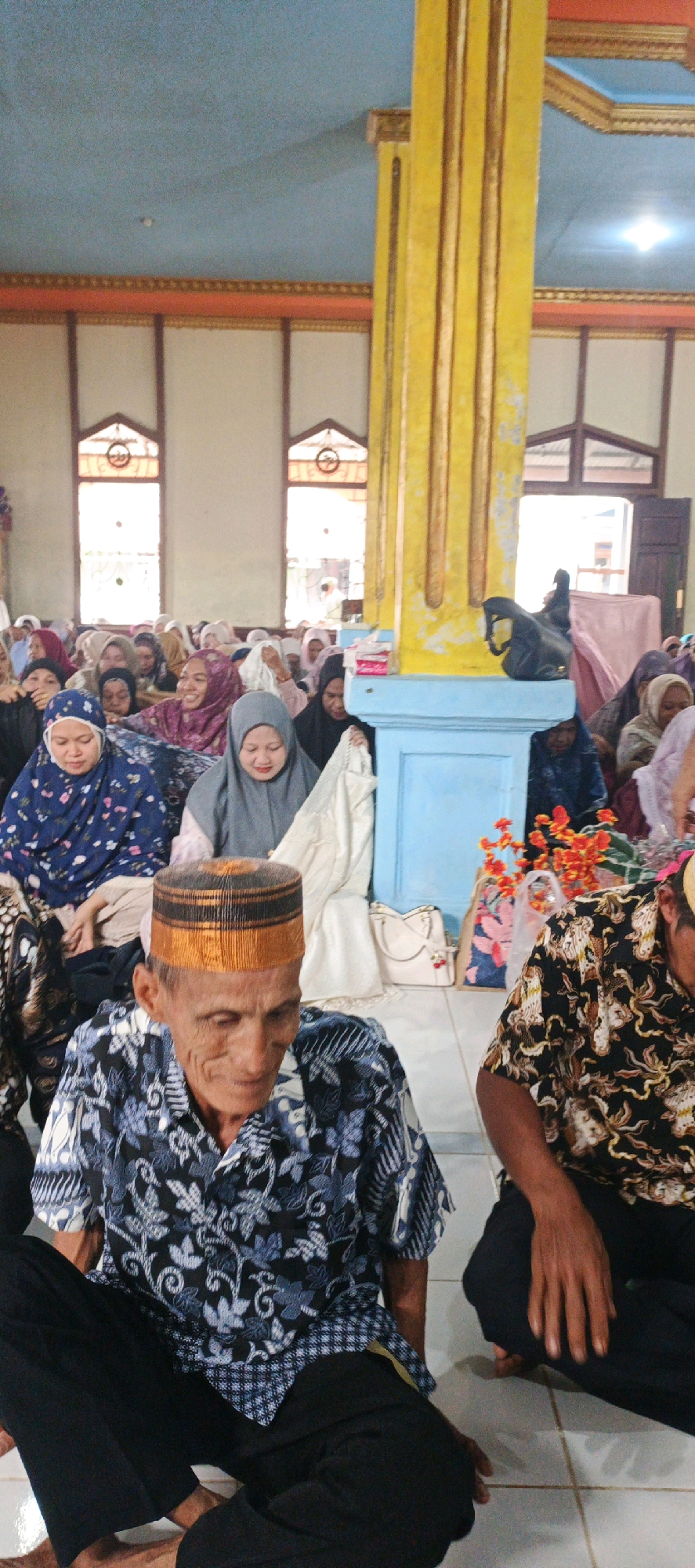 In Simbur Naik, Indonesia, a group of people are gathered indoors, likely for a religious or community event. The room features a pale blue ceiling with decorative orange and gold trim along the walls and ceiling. A prominent, tall, yellow pillar with gold vertical accents stands to the right of the frame. The floor is tiled with off-white squares.
In the foreground, two men are seated on the floor. The man on the left is older, wearing a patterned blue and white shirt and a striped cap with orange and black colors. He is looking down. To his right, another man, partially visible, wears a patterned black and gold shirt and is also seated.
Behind the men, a crowd of people, predominantly women wearing hijabs, are seated on the floor. Some women are looking towards the front, while others are interacting amongst themselves. A woman in a grey hijab is visible in the middle ground, and another in a blue and white patterned hijab is to her left. A light-colored blanket or cloth is draped over some individuals. In the background, two windows with arched tops are visible, letting in natural light. Several bags, including a white tote bag and a patterned tote bag with red flowers, are placed near the pillar. The overall atmosphere appears serene and communal.