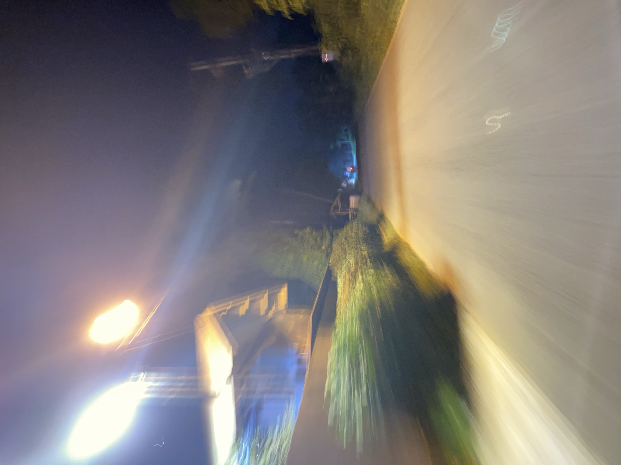 This media file is a photograph taken at night, likely during nighttime or early morning hours given the artificial lighting and dark sky. The image depicts a street scene with a road on the right side, appearing to be paved and illuminated by a streetlamp. The left side of the image shows trees and some structures, possibly houses or buildings. The overall impression is one of motion blur, suggesting the photograph was taken from a moving vehicle or with a shaky hand. The lighting is a mix of the yellow glow from a streetlamp and cooler blueish tones from distant lights, possibly from homes or other sources. The trees are illuminated from below, casting long shadows and creating streaks of light.
There are no discernible people in the image. Objects visible include streetlights, trees, and hints of structures. The scene is an outdoor, suburban or semi-rural environment with a road, vegetation, and some man-made structures. The time of day is clearly night, and the weather appears clear due to the visibility of the sky and lights.
Notable details include the strong motion blur which gives the image an abstract and dynamic quality. The presence of streetlights and illuminated trees suggests a residential area. The low angle and perspective create a sense of looking down a street.
There is no visible text in the image. The emotional tone conveyed is somewhat mysterious and perhaps a little unsettling due to the darkness and motion blur, but also potentially peaceful due to the quiet street scene. The location context of Lhokseumawe, Indonesia, does not provide specific visual cues within the image itself.