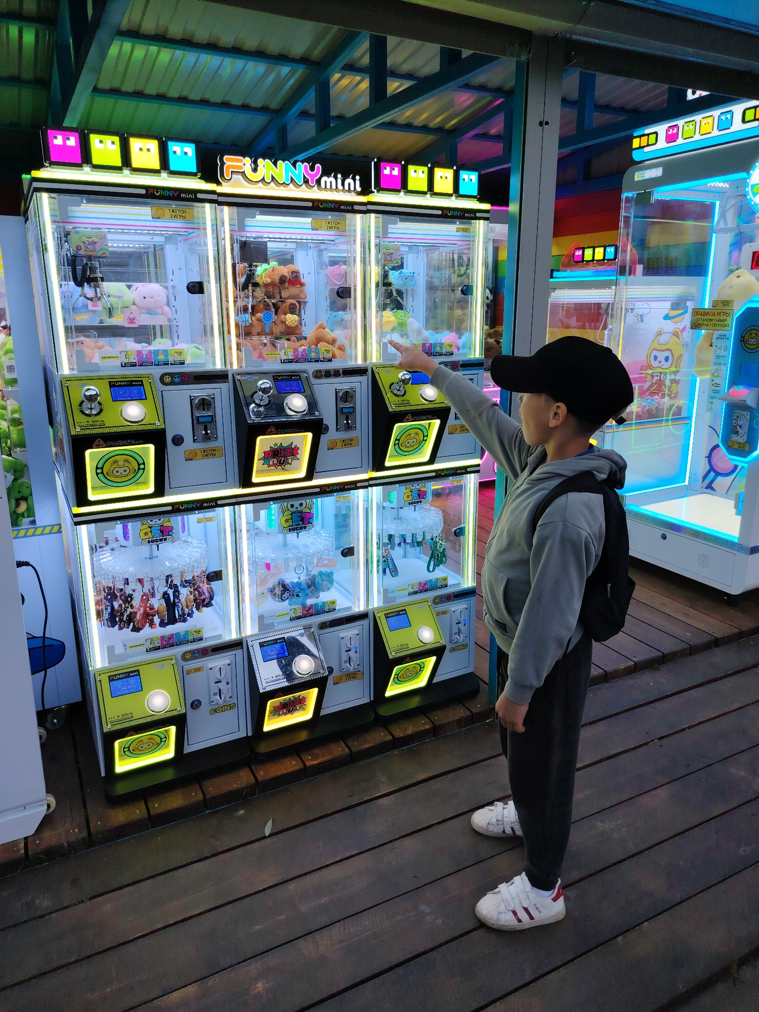 A young boy in a gray hoodie, dark sweatpants, white sneakers with red stripes, and a black cap is standing in front of a three-tiered "FUNNY mini" claw machine arcade game, pointing his right index finger towards the middle game on the top tier. He has a black backpack on his back. The claw machines are brightly lit with various prizes inside, including stuffed animals and figurines. The machines are labeled with text in Russian. The arcade game is located in Chaikovsky, Russia. The floor is made of wooden planks. Another arcade game can be seen to the right. The ceiling above is constructed with a blue and green metal frame and is partially visible.