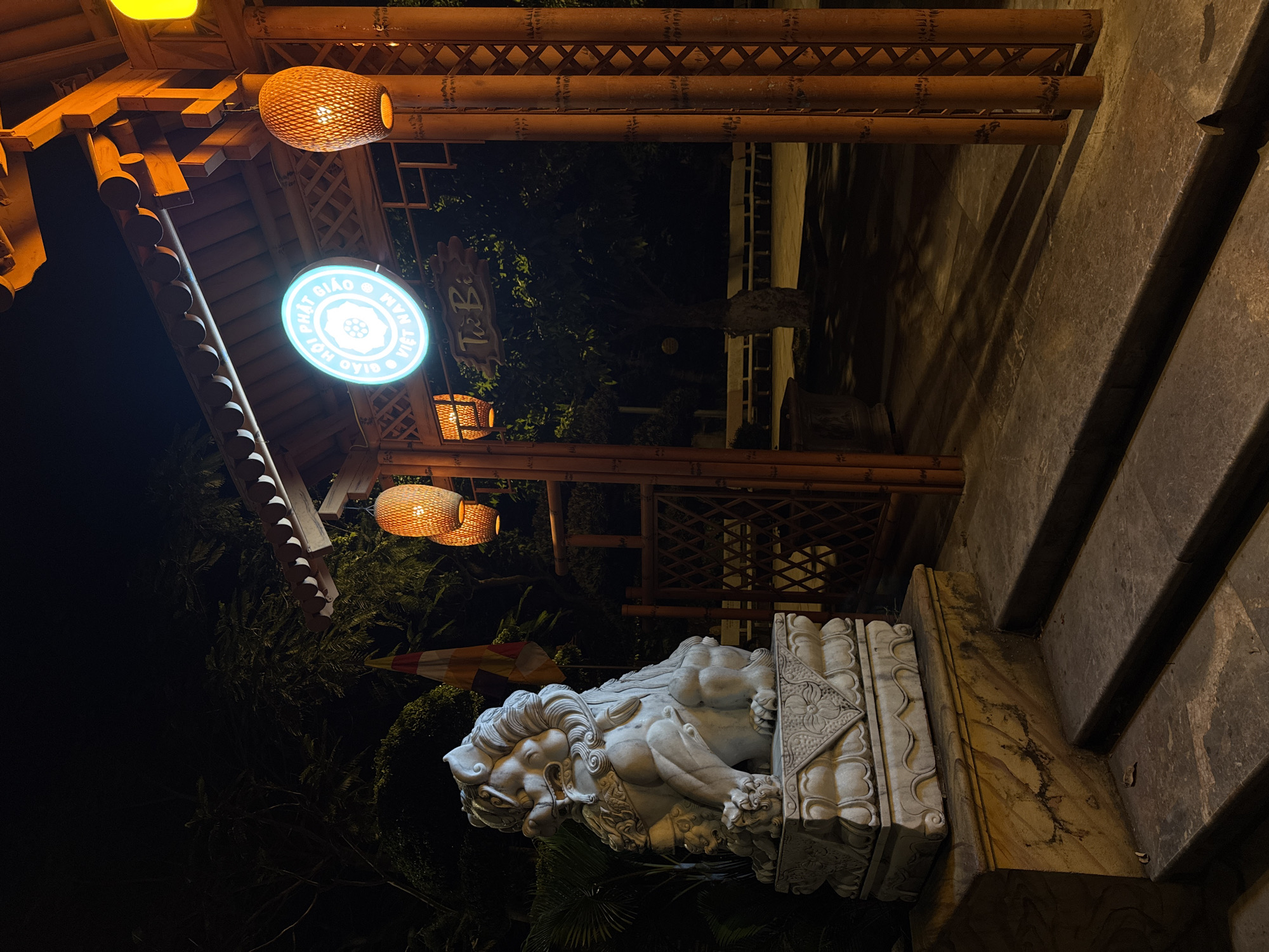 The image depicts the entrance to Tu Bi Buddhist temple at night in Da Nang, Vietnam. A stone statue of a mythical lion-dog, known as a 'foo dog', guards the left side of the entrance, positioned on a stone pedestal. The entrance itself is an arched gateway constructed from bamboo, illuminated by warm light from hanging lanterns. A circular sign with blue neon lettering is visible above the entrance. The ground leading up to the entrance consists of stone steps. Benches are visible in the background, suggesting a seating area within the temple grounds. The overall scene is dimly lit, with shadows cast by the structures and vegetation.