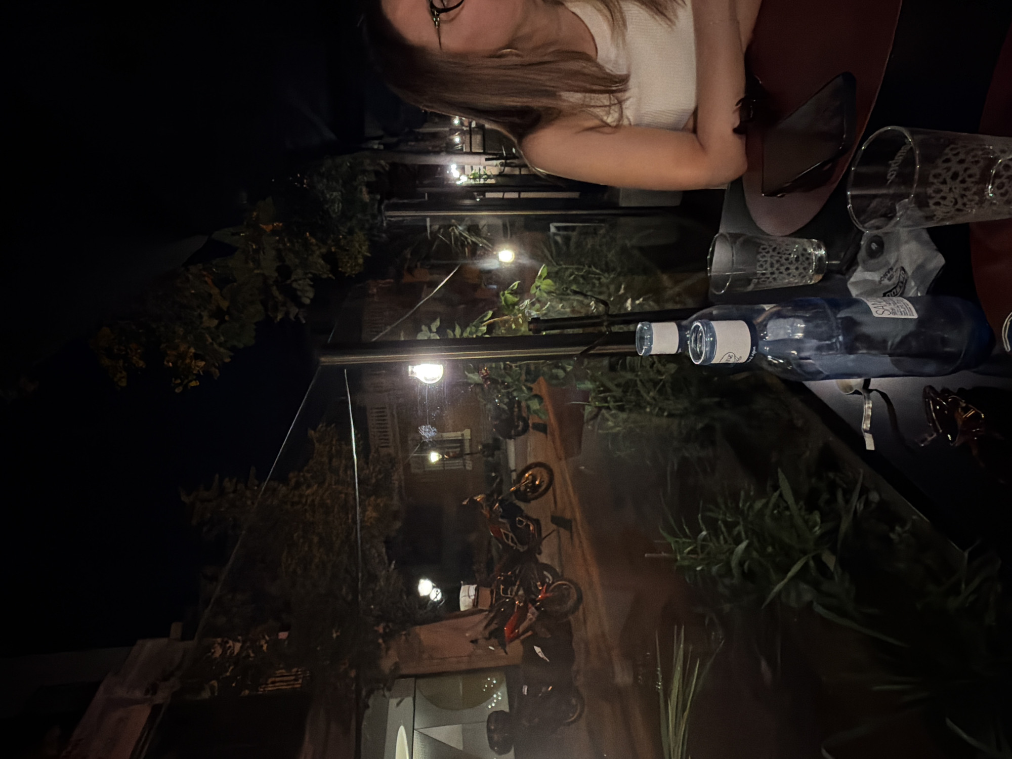 A nighttime outdoor scene shows a woman sitting at a table in what appears to be a restaurant patio in Unknown, Unknown. The table setting includes two glasses with animal print designs, a bottle of water, a brown plate, and a pair of sunglasses. Outside the restaurant, several motorcycles are parked on the street. String lights are visible along the patio, and vegetation is present, providing a natural backdrop.
