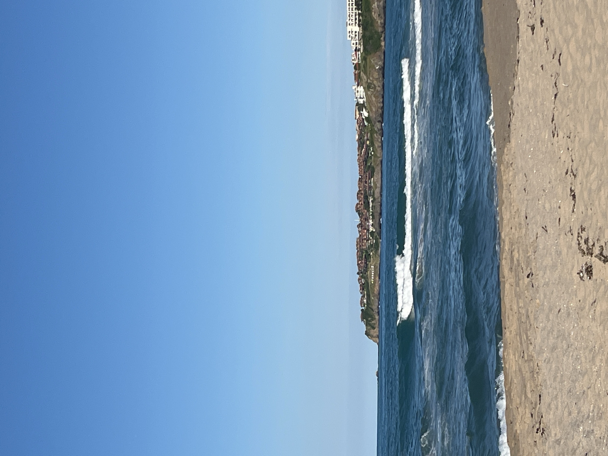 The image captures a coastal scene in Sozopol, Bulgaria, on a clear, sunny day. The sky is a vibrant, cloudless blue, dominating the upper half of the frame. Below, the deep blue sea is active with moderate waves that break into white foam as they approach the shore. The water exhibits shades of teal and turquoise, suggesting clarity.
In the distance, a prominent cliffside overlooks the sea, densely populated with buildings. These structures feature terracotta roofs and a mix of white and earth-toned facades, giving the impression of a historic town or resort. The cliff is green with vegetation.
The foreground is a sandy beach, appearing somewhat coarse and scattered with small pieces of debris, possibly dried seaweed or twigs. The sand transitions from a lighter shade at the very bottom of the image to a slightly darker tone where it meets the water's edge. There are no people visible in the image, and the focus is entirely on the natural landscape and the coastal settlement. The overall impression is one of a tranquil, picturesque seaside location.
