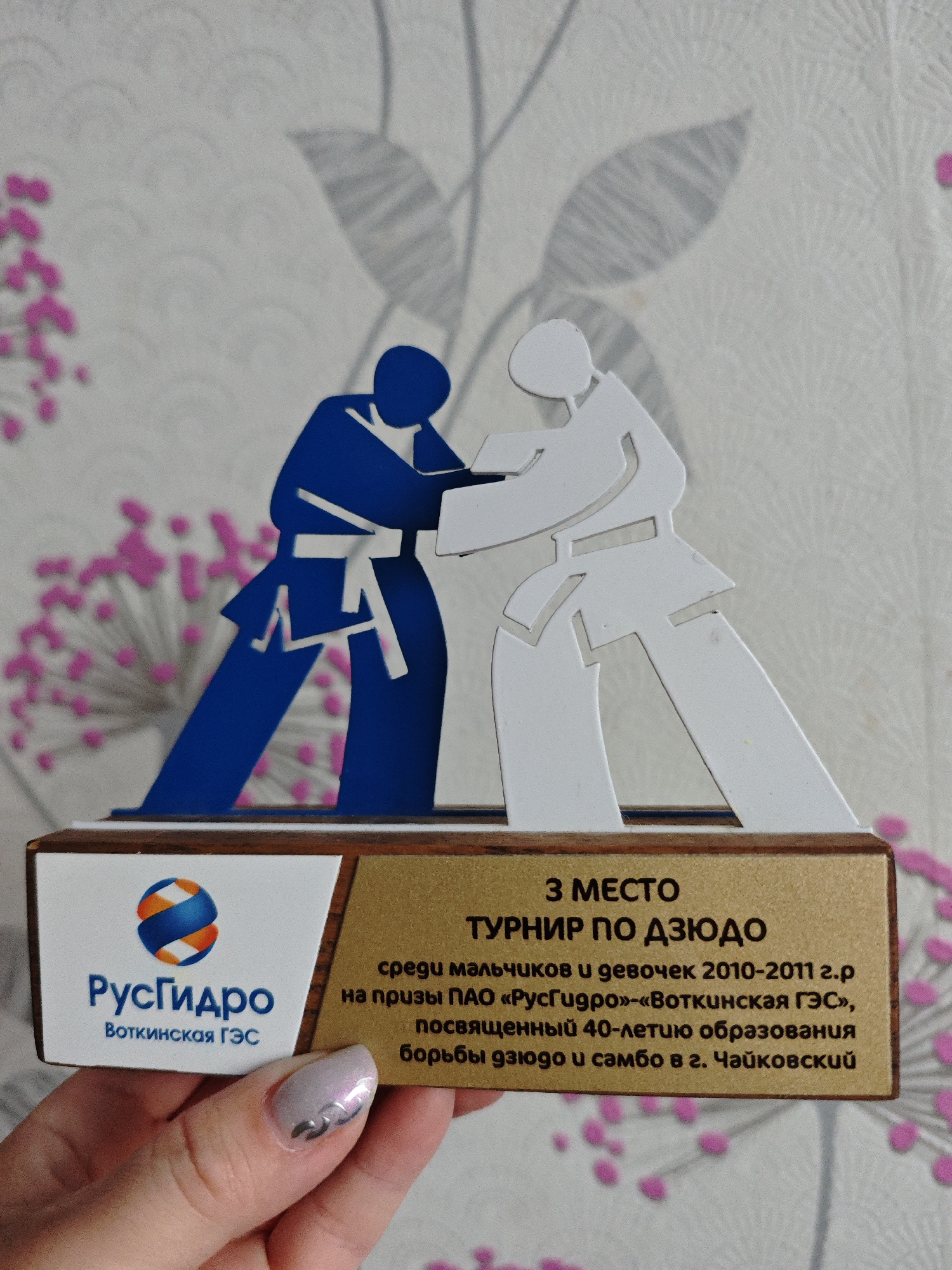 The image shows a judo tournament award held by a hand with a light-colored nail. The award is comprised of a wooden base with a white label on the left side bearing the logo of "РусГидро" (RusHydro) and the text "Воткинская ГЭС" (Votkinsk GЭС). Next to this section is a gold label that reads "3 МЕСТО" (3rd place), "ТУРНИР ПО ДЗЮДО" (Judo Tournament), "среди мальчиков и девочек 2010-2011 г.р" (among boys and girls 2010-2011), "на призы ПАО «РусГидро»-«Воткинская ГЭС»" (at the prizes of PJSC "RusHydro" - "Votkinsk GЭС"), "посвященный 40-летию образования борьбы дзюдо и самбо в г. Чайковский" (dedicated to the 40th anniversary of judo and sambo wrestling education in the city of Chaikovsky). Atop the base are two figures in judo poses, one in blue and the other in white. The background is a wall with a floral design. The city of Markovskii is not mentioned in the text.