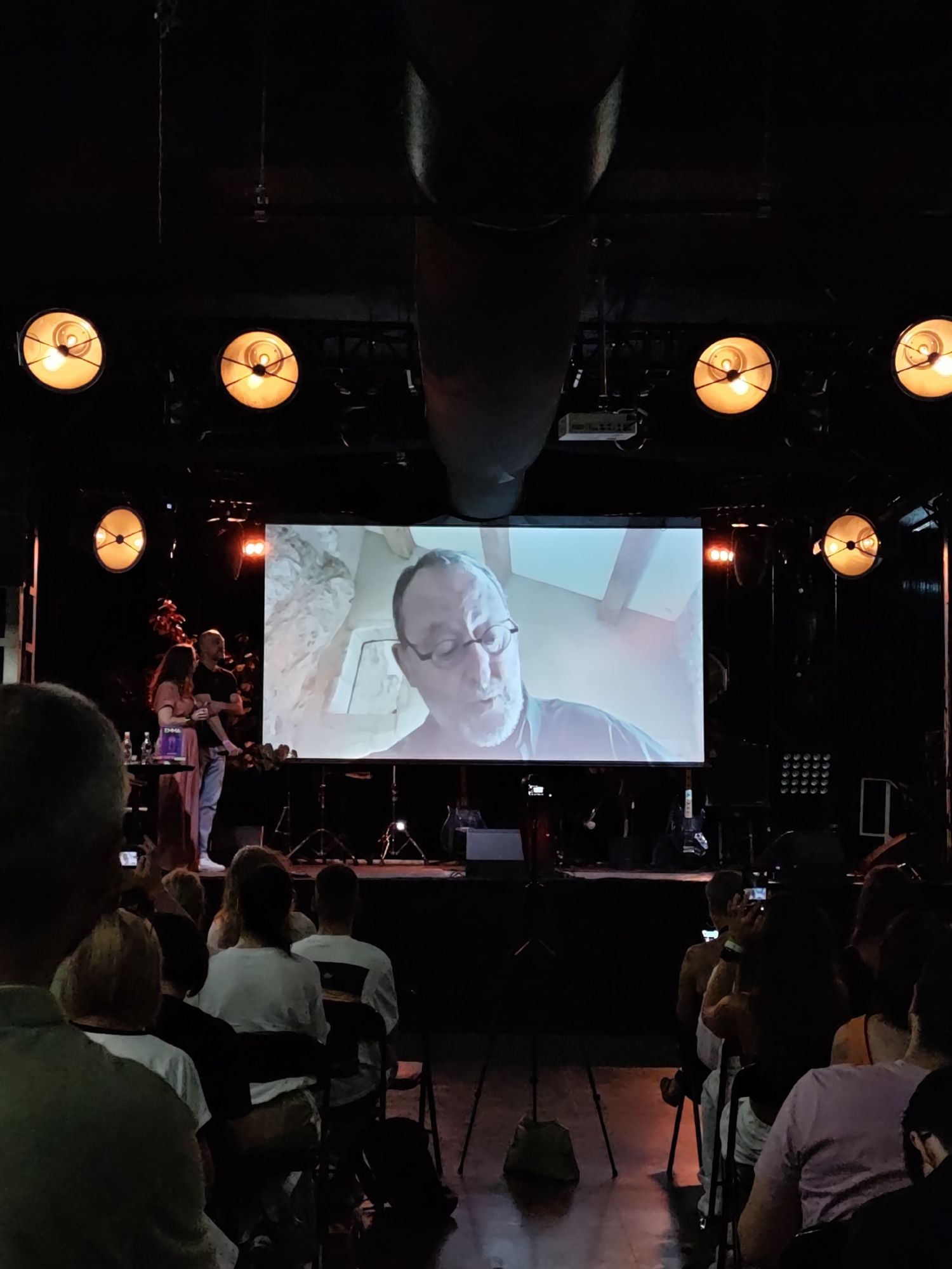 The image depicts an indoor event, likely a presentation or performance, in Lviv, Ukraine. A large screen displays a man with glasses speaking, positioned in front of a textured background. On a stage below the screen, two individuals, a woman in a long pink dress and a man in a dark t-shirt and jeans holding a baby, stand near a table with bottles and a book. The venue appears to be a dark room with a stage and an audience seated in chairs. Several spotlights are visible above the stage, casting orange light. The audience members are mostly facing the screen, some are holding cameras or phones. The overall atmosphere suggests a public gathering focused on the projected content.