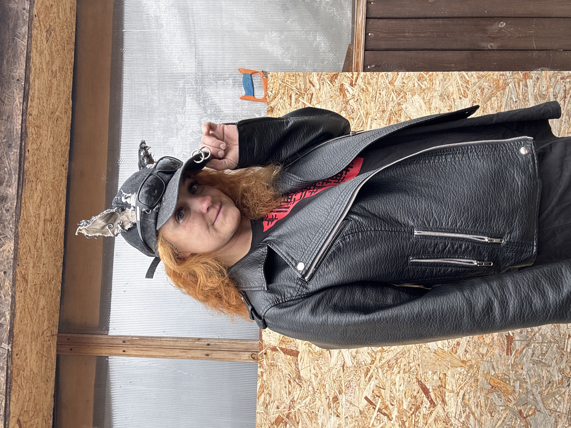 A woman with reddish-orange hair wearing a black leather jacket, a black t-shirt, and a grey baseball cap with metallic accessories is posing for a photo. The woman is holding up her left hand with the fingers slightly bent. She is standing in front of a building that appears to have a corrugated, translucent wall panel and a wooden exterior. In the background, there is a wooden structure and a small handsaw is visible. The location is Chaikovsky, Russia.