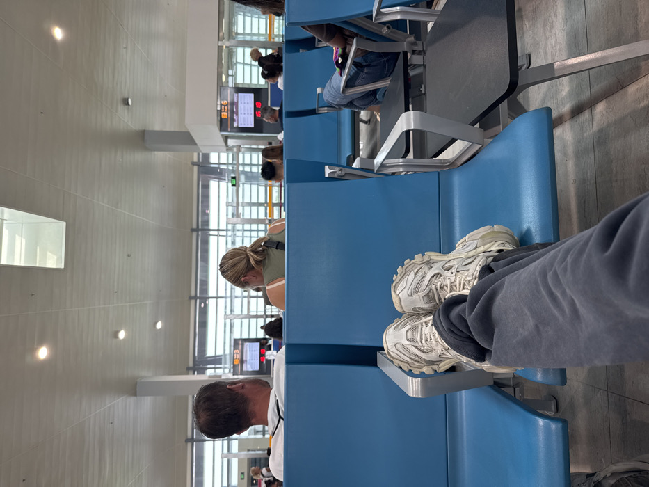 This image depicts a waiting area within the Abeid Amani Karume International Airport in Zanzibar City, Tanzania. The foreground shows the observer's legs, clad in dark grey sweatpants and white chunky athletic shoes, resting on a light blue airport seat. To the left, a man with short brown hair in a white t-shirt and a woman with blonde hair in a ponytail wearing a green tank top are seated on identical blue chairs, both facing away from the camera. Several other passengers are partially visible further down the seating rows. In the background, large windows overlook what appears to be an outdoor airport area, and gate signs for "Gate 3" and "Gate 6" are clearly visible, displaying digital information. The ceiling is white with integrated linear lighting and rectangular skylights, indicative of a modern airport terminal.