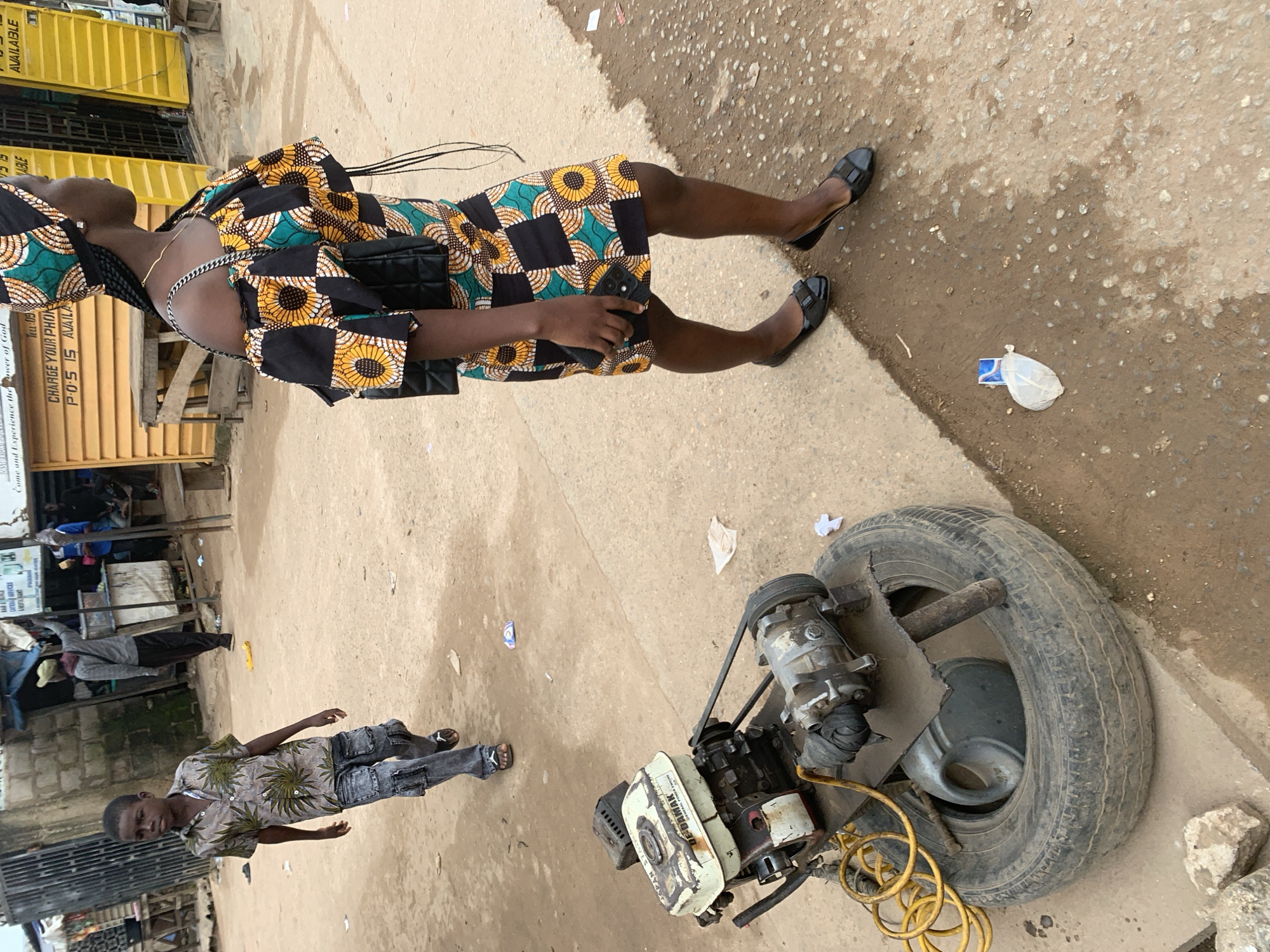 The image captures a scene on a dirt road in Shagamu, Nigeria, with a focus on a young boy and a woman. The boy, walking from left to right, wears a short-sleeved patterned shirt and distressed denim cargo pants. In the foreground, a portable generator is mounted on a tire, with yellow tubing coiled nearby. To the right, a woman, seen from the waist down, is walking away from the camera. She is wearing a vibrant, patterned dress with off-the-shoulder sleeves and a matching headwrap. She carries a black purse and is wearing black ballet flats. The background shows a street environment with buildings, stalls, and other pedestrians, suggesting a commercial or public area. The ground is a mix of dirt and paved surfaces, with scattered debris.