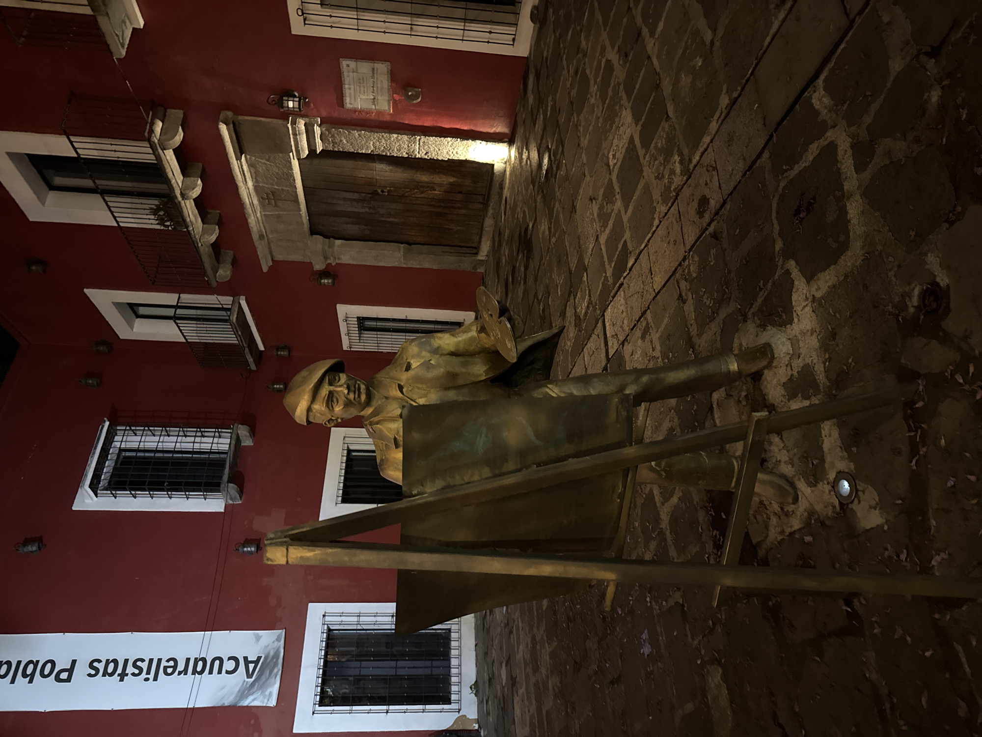 A bronze statue of an artist stands on a cobblestone street in Puebla, Mexico. The artist wears a beret and holds a palette, positioned in front of an easel. The building behind the statue is painted red and features white-framed windows with black metal bars, along with a wooden door illuminated by a light above. A banner with Spanish text, "Acuarelistas Puebla," hangs on the building. The cobblestone street has a slightly upward incline and a light embedded in the stones near the statue.