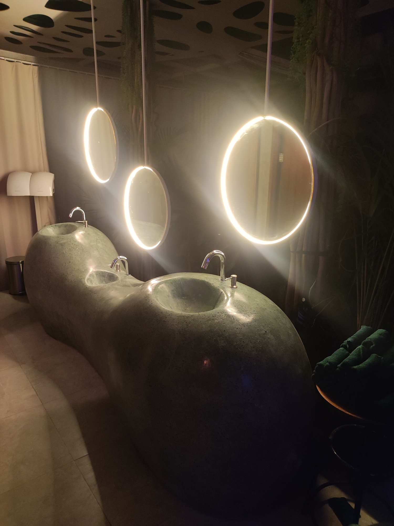The image shows a modern bathroom with a unique, flowing sink structure. The sink is made of a dark, textured material and appears to have multiple basin-like depressions, each with a chrome faucet. Above the sink, three circular mirrors with integrated halo lighting are suspended from the ceiling. The ceiling itself has a decorative pattern of irregular dark shapes against a lighter background.
To the left of the sink, there are two white hand dryers mounted on a beige wall, and a cylindrical silver trash can. Behind the sinks, a dark wall is visible, with some greenery and what appears to be decorative tree-like elements. On the right side of the image, a stack of folded green towels is placed on a circular surface, next to a dark object, possibly a foot bath.
The overall aesthetic is contemporary and somewhat organic, with soft lighting from the circular mirrors creating a warm glow. The floor appears to be tiled. This scene is likely located in a public restroom, possibly in a hotel, spa, or restaurant in North Kuta, Indonesia, given the characteristic tropical elements and modern design often found in such establishments in the region.