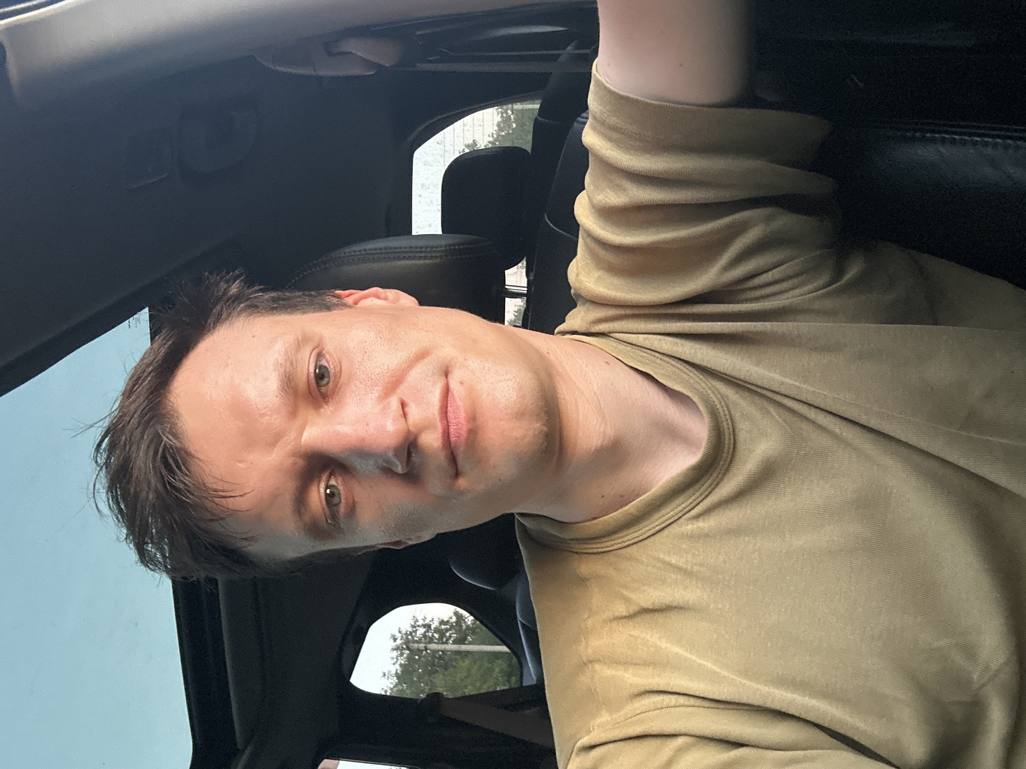 A selfie taken from inside a car shows a man with short, dark, messy hair. He has fair skin, light-colored eyes, and a neutral expression. He is wearing a light brown t-shirt. The car has a sunroof, which is open and lets in a bright sky. The interior of the car is dark, with leather seats visible. Outside the car, through the windows, green trees and a bright sky can be seen. The car is likely parked or driving on a road in Makiv, Ukraine.