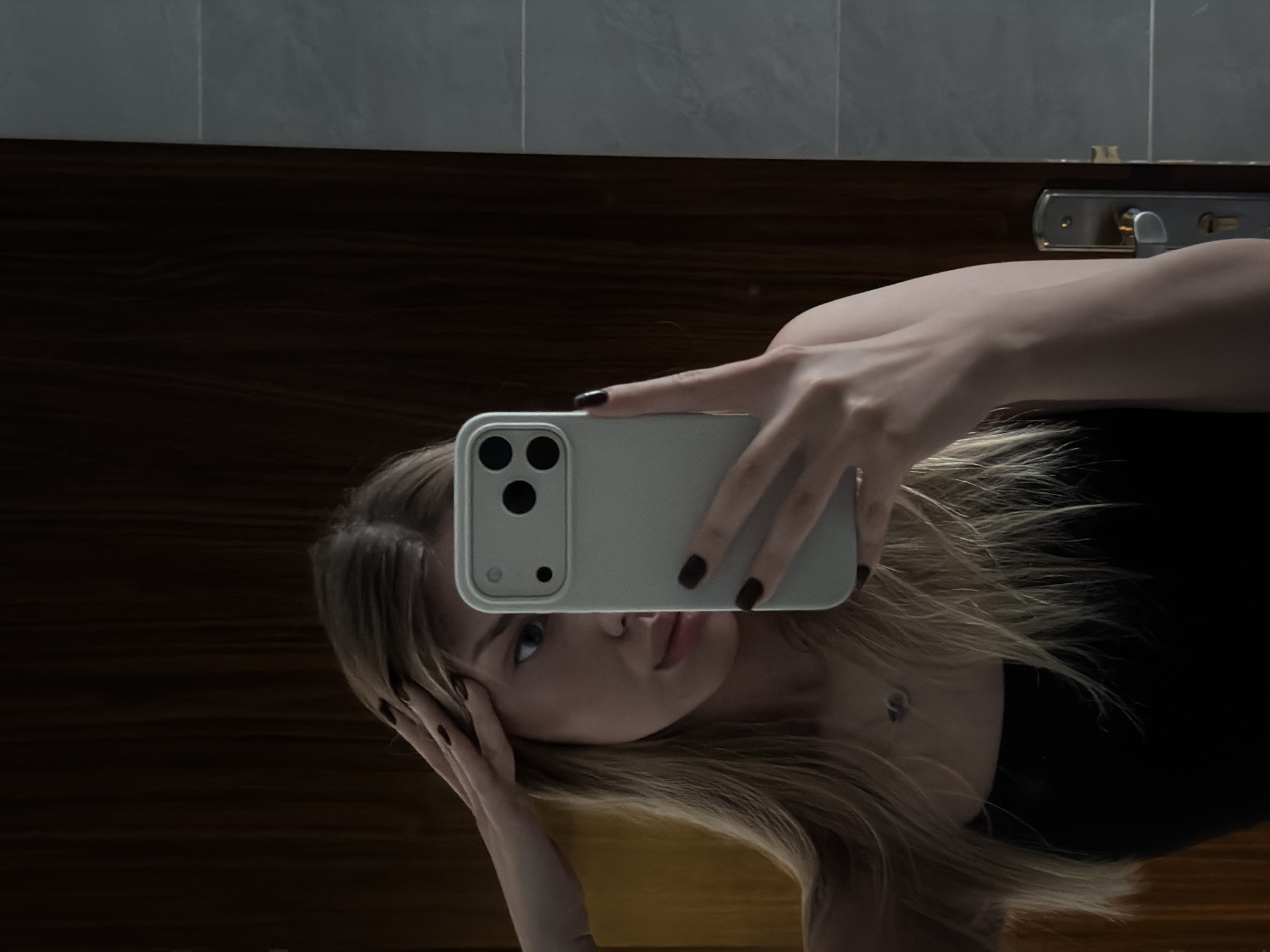 This media file shows a selfie taken by a young woman in a bathroom. She is holding a light-colored smartphone in her right hand, angled to capture her reflection in a mirror. The woman has blonde hair, styled loosely around her face, and is wearing a black top. Her left hand is resting on her head, and her expression appears thoughtful or slightly melancholic.
The background suggests a modern bathroom setting. A dark wooden vanity or door is visible behind her, with a stainless steel door handle and lock assembly mounted on it. Above the wood paneling, there are tiled surfaces, possibly the wall or a backsplash. The lighting is dim, creating shadows and highlighting the woman's face and hand. The overall mood is intimate and personal.
There is no visible text in the image. The time of day is not explicitly indicated, but the dim lighting might suggest evening or a room with limited natural light. The location context is Đà Nẵng, Vietnam, but there are no specific visual cues in the image to confirm this. The activity is the act of taking a selfie, a common practice for personal documentation or social media sharing.
