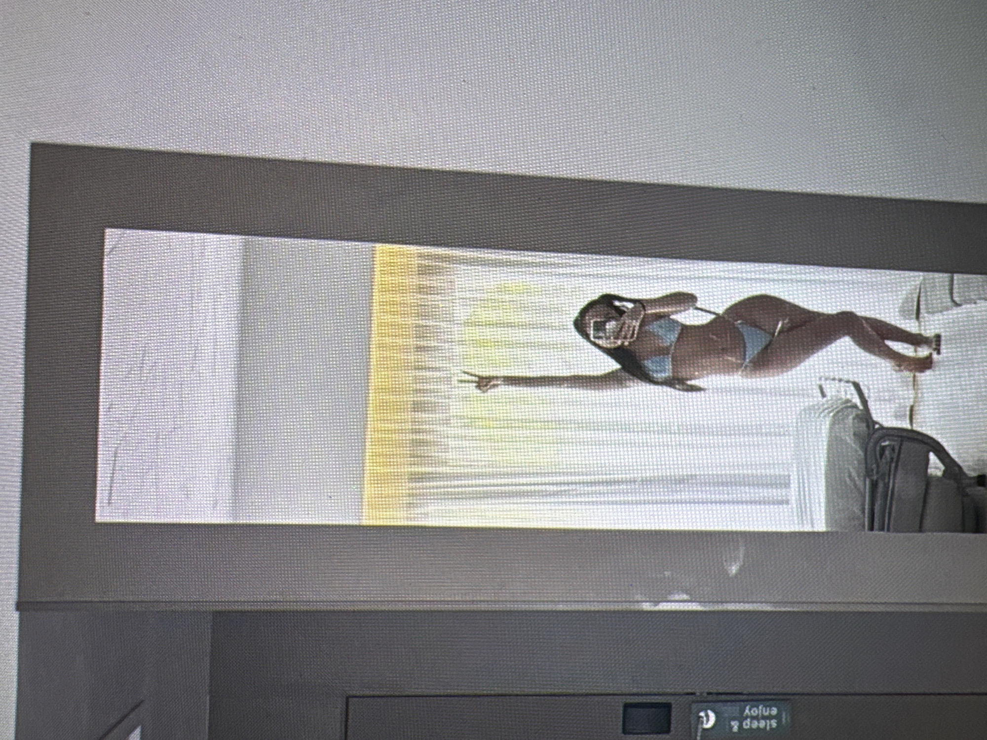A full-length mirror reflects a person in a light blue bikini standing in a room with white curtains and yellow lighting. The person is holding up a hand in a peace sign and looking at their reflection. To the left of the mirror, a dark gray door with a black rectangular handle is visible, and a label reading "sleep & enjoy" is attached to it. A bed with white bedding is partially visible in the reflection, positioned behind the person. The environment appears to be a hotel room or a similar lodging, likely in Castelo Branco, Portugal, given the context.