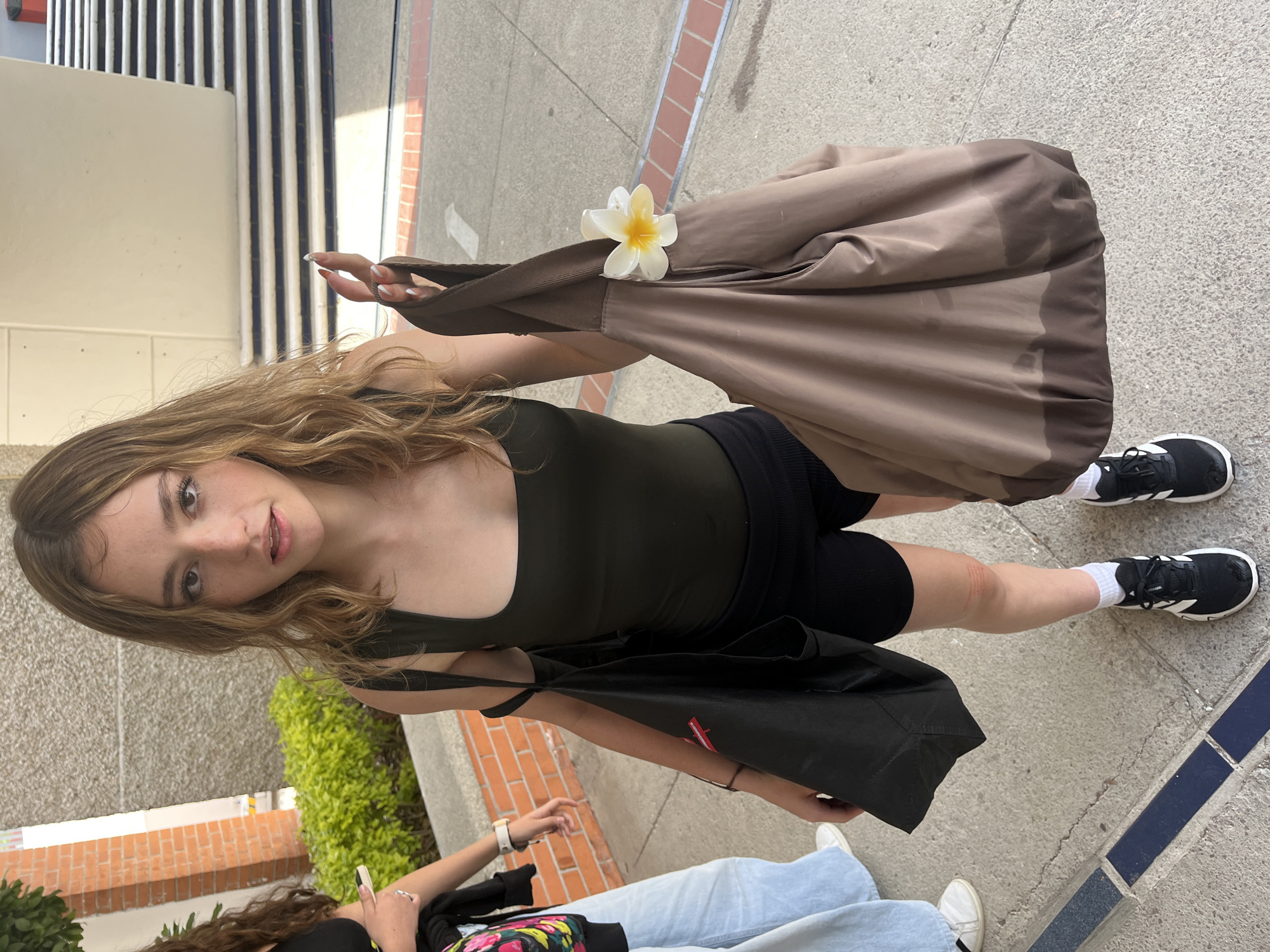 The image captures a young woman standing outdoors in what appears to be an urban or campus setting in Puebla, Mexico. She is the central subject, looking directly at the camera with a calm, neutral expression. Her light brown, wavy hair cascades over her shoulders. She is wearing a dark olive green tank top, black biker shorts, black Adidas sneakers with white soles, and white socks.
In her right hand, she holds a large, soft brown tote bag, from which a vibrant white and yellow plumeria flower is visibly tucked into the strap. A black tote bag is carried over her left shoulder. The ground beneath her is a light grey concrete, accented with dark blue and brick-colored tiled borders.
The background includes a building with distinctive white and dark vertical stripes, possibly indicating stairs or an architectural facade, and a concrete wall. Green foliage and a low brick wall are also present, adding to the outdoor ambiance.
Partially visible in the lower foreground is another person, likely female, wearing light blue jeans, white shoes, and a black top adorned with colorful floral patterns. This person appears to be holding or looking at a mobile phone.
The scene is well-lit by natural sunlight, suggesting a clear day. The plumeria flower is a notable detail, aligning with the warm climate often associated with Mexico. Aside from the Adidas logo on the main subject's shoe, no other discernible text is visible. The overall impression is a casual, everyday moment captured in a bright, inviting environment.