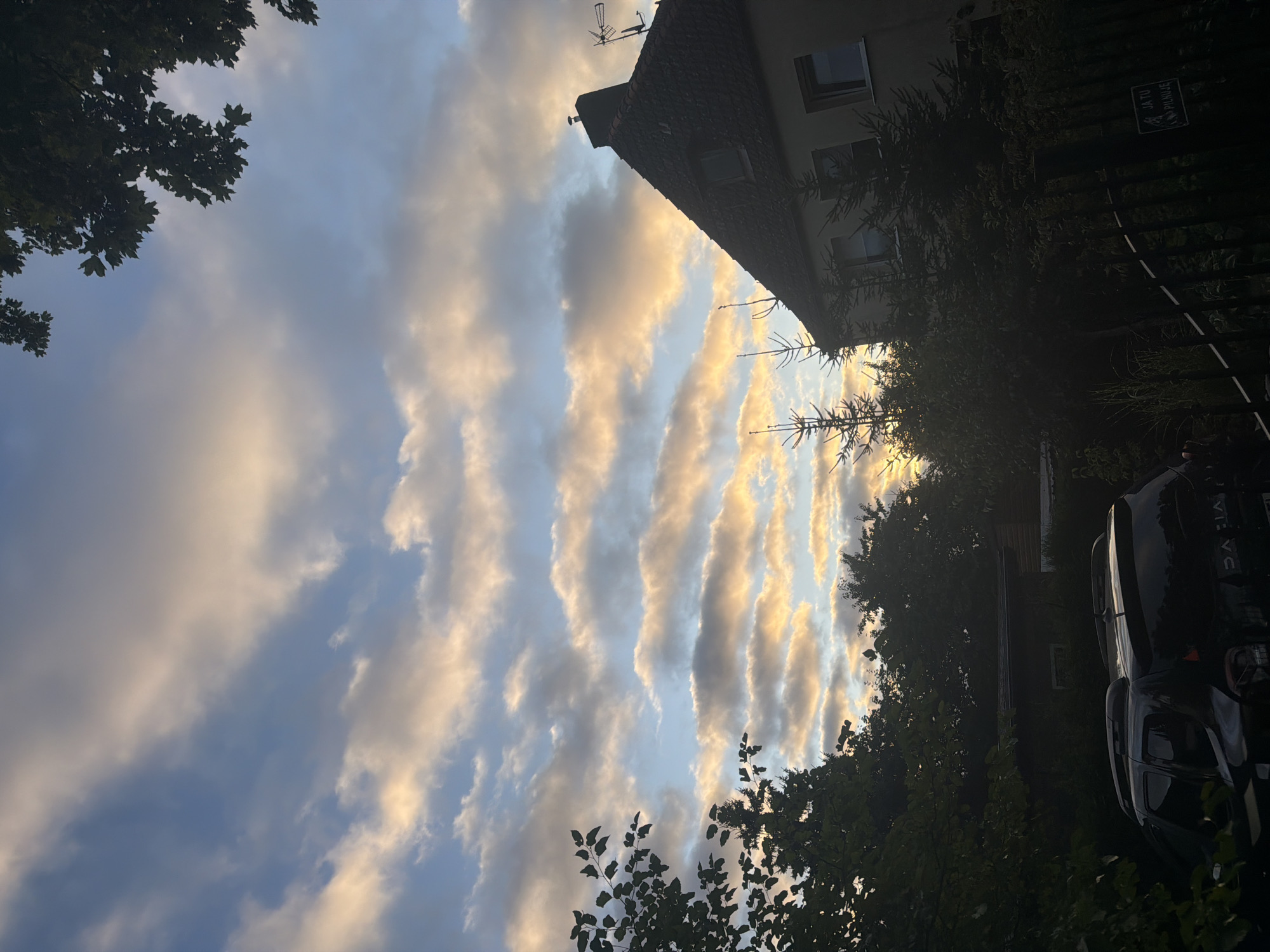 The image depicts a residential area in Szczecin, Poland, under a sky filled with dramatic clouds illuminated by the setting sun. In the foreground, a black SUV is parked, partially obscured by the shadows of surrounding trees and foliage. To the right, a fence with a small sign is visible, and beyond it, a building with a dark-tiled roof and windows stands under the colorful sky. The overall scene captures a serene, late-day atmosphere in a suburban or semi-rural setting within the city.