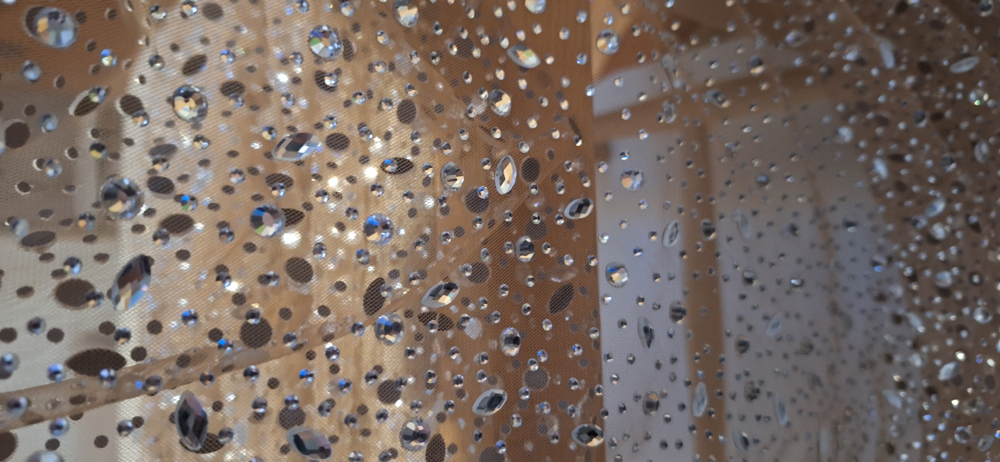 The image shows a close-up of a shimmering fabric. The fabric appears to be a light tan or beige color and is covered in a pattern of small, reflective rhinestones and darker circular perforations. The rhinestones vary in size and shape, with some appearing round and others more elongated or faceted. The perforations also vary in size, adding to the textured and patterned look of the fabric. The image is possibly taken in an indoor setting with indirect lighting, creating a diffused glow and highlights on the rhinestones. The fabric's draping suggests it is suspended or hanging. The location is not discernible from the image.