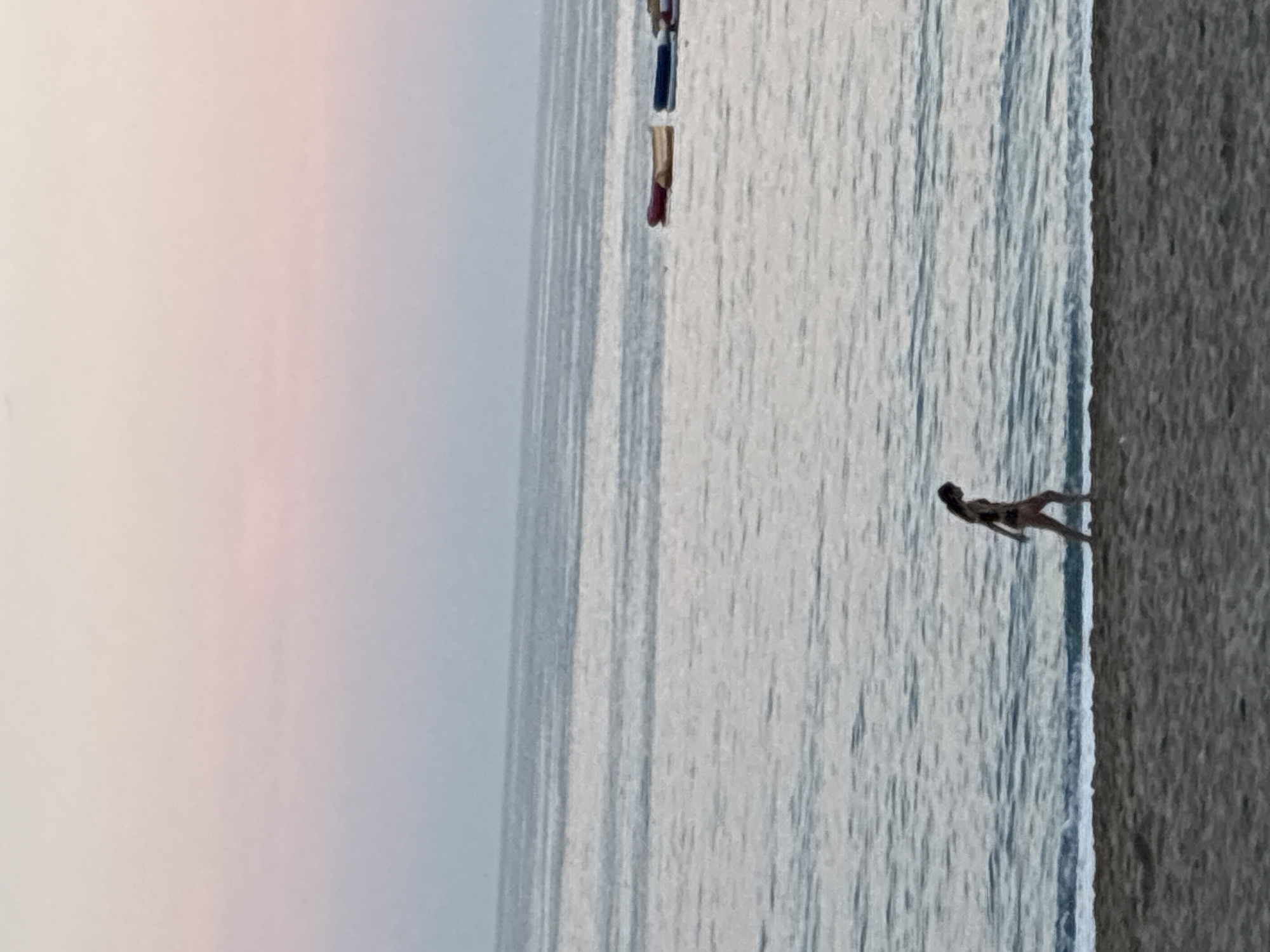 The image displays a tranquil coastal scene in Alanya, Turkey, under soft, diffused lighting, likely during dawn or dusk. The sky occupies the upper portion, exhibiting a gentle gradient from pale beige-pink at the top to a muted blue-gray near the horizon. The mid-ground is dominated by the calm, expansive sea, rendered in shades of light blue-gray with subtle surface ripples. In the middle-right distance on the water, several small, rectangular objects, appearing to be rafts or small boats, are visible in colors including dark red, light yellow, and dark blue. The foreground consists of a sandy beach, presented in subdued brown and gray tones, showing fine textural details. A lone female figure walks along the waterline from left to right, depicted in profile, wearing a dark-colored swimsuit. Gentle waves break at the immediate shoreline, creating a thin line of white foam where the water meets the sand. The overall atmosphere is serene and understated.