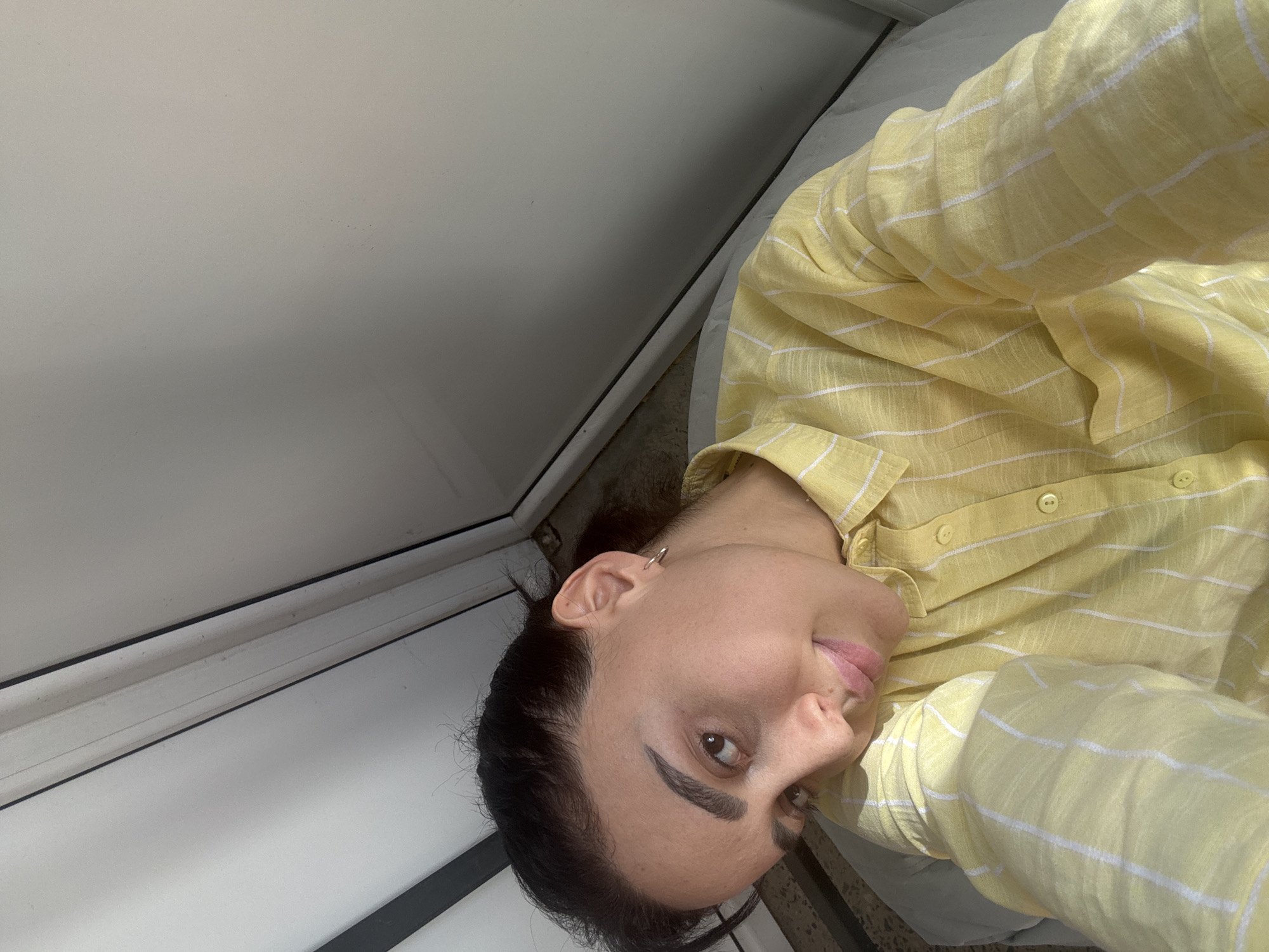 A woman is indoors, possibly in a building in Tbilisi, Georgia. The image is taken from a low angle, showing her head and upper body. She is wearing a light yellow shirt with white stripes. She has dark hair and is looking towards the camera. A white wall is visible in the background to the left.